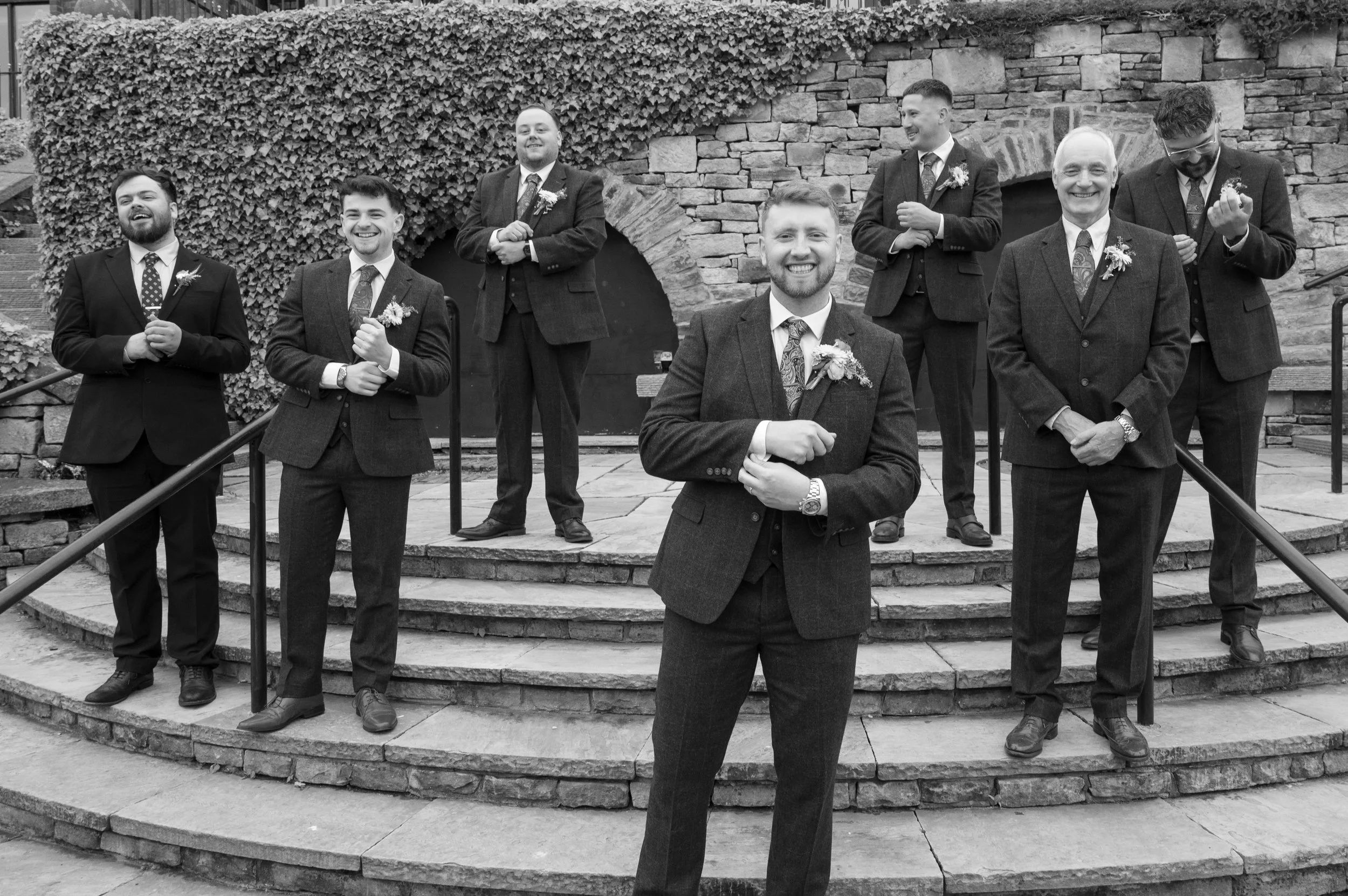A group of eight men dressed in formal suits with boutonnières, standing on stone steps in front of a brick and ivy wall, smiling and posing for a photo.