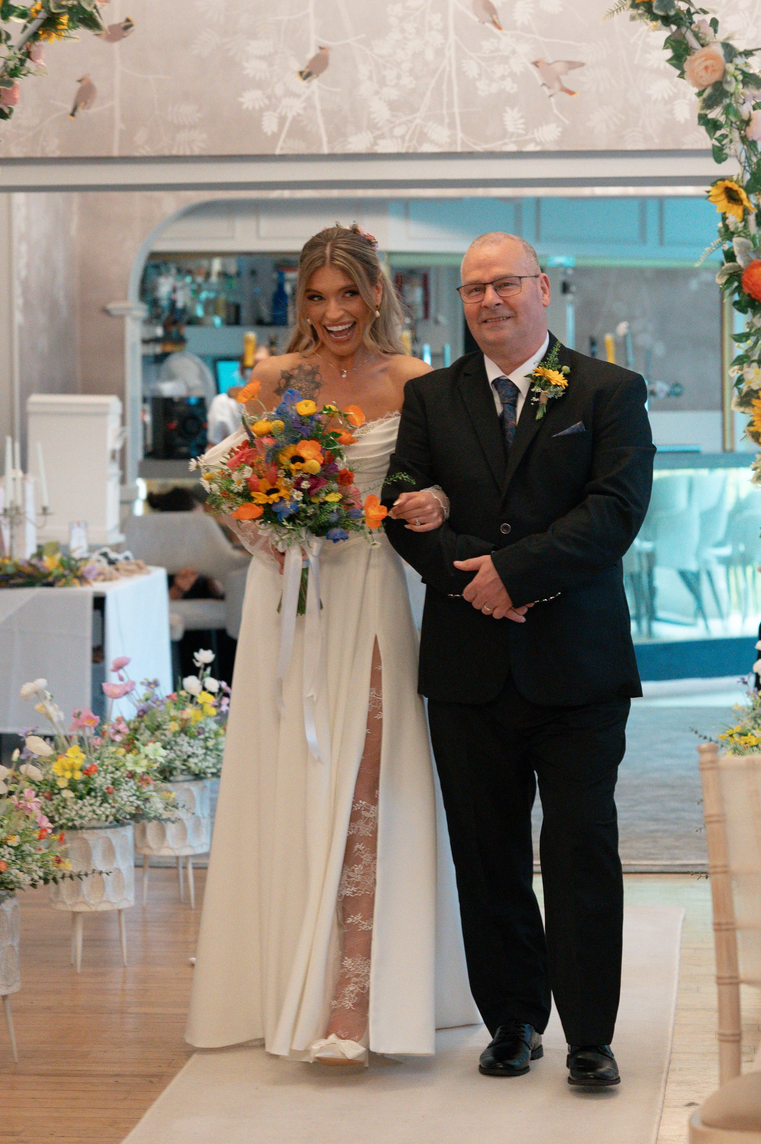 A bride holding a colorful bouquet of flowers and smiling as she walks down the aisle with a man in a black suit. The area is decorated with flowers and a floral archway.