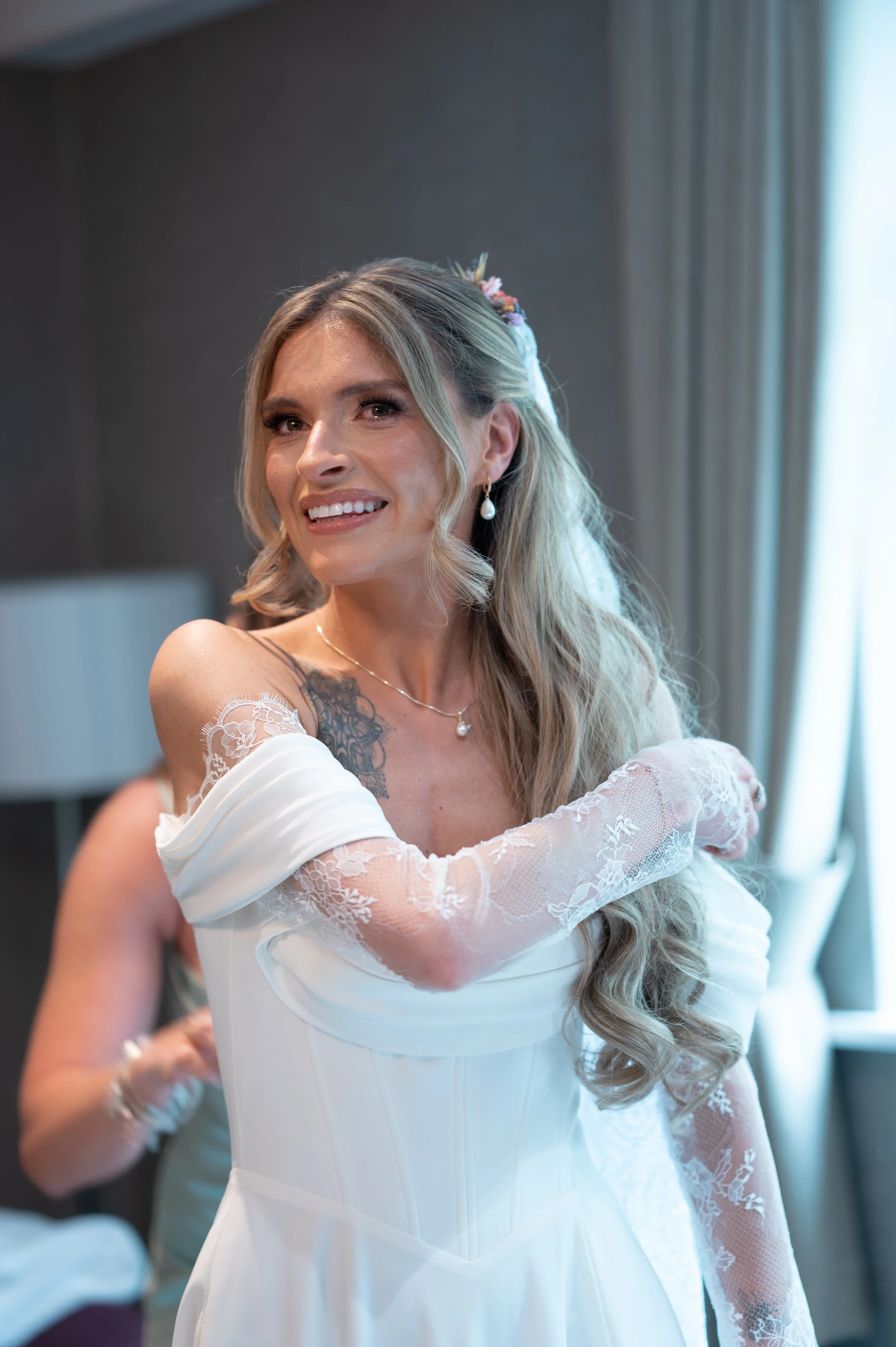 A bride in a white wedding dress and lace veil smiling and embracing, with a woman in the background helping with her dress. The bride has long, wavy hair with floral accessories and pearl earrings, standing near a window with curtains.