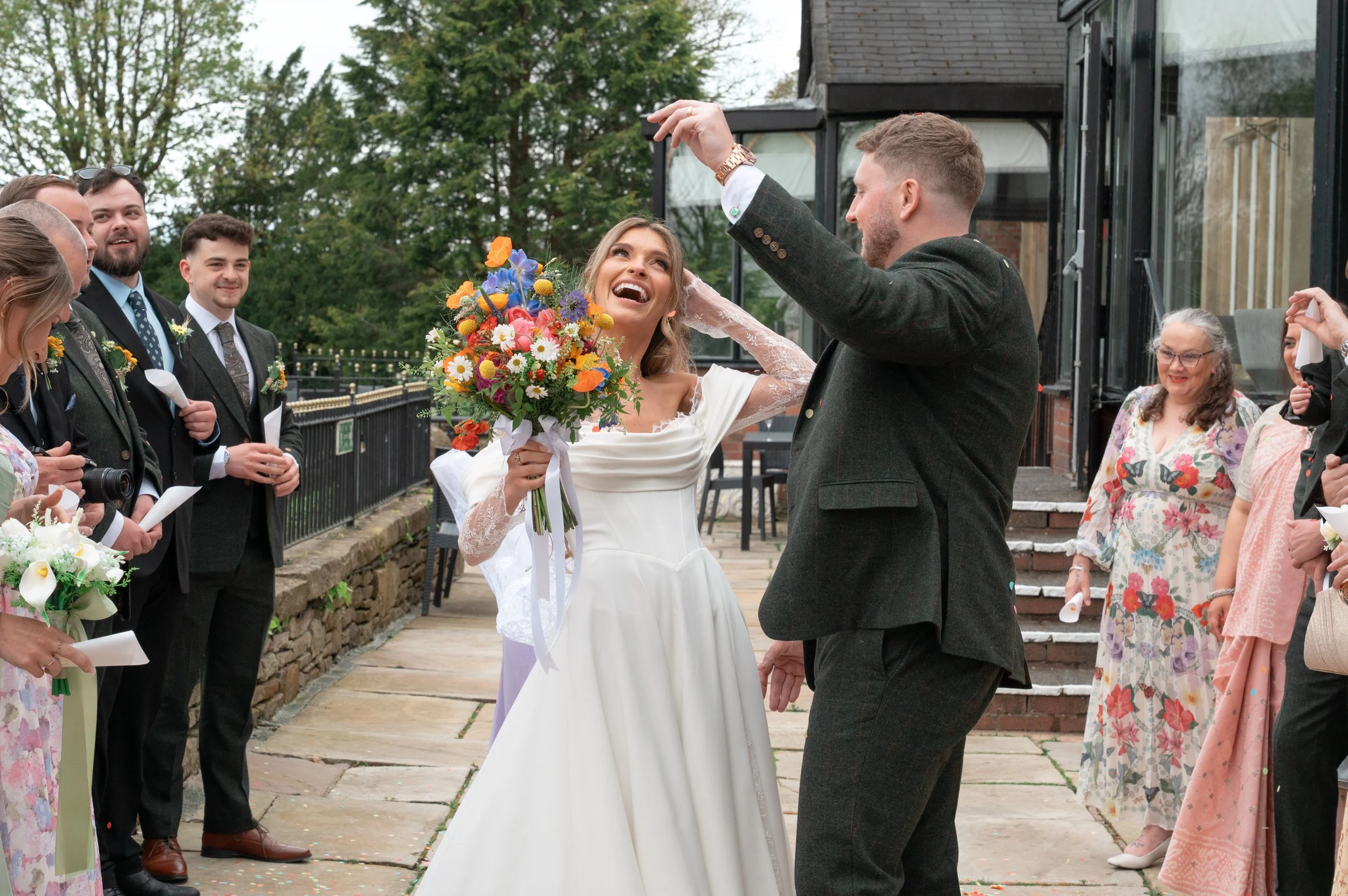 A bride and groom holding hands during their outdoor wedding ceremony, surrounded by friends and family, with the bride smiling as the groom holds her hand and a colorful bouquet in her other hand.