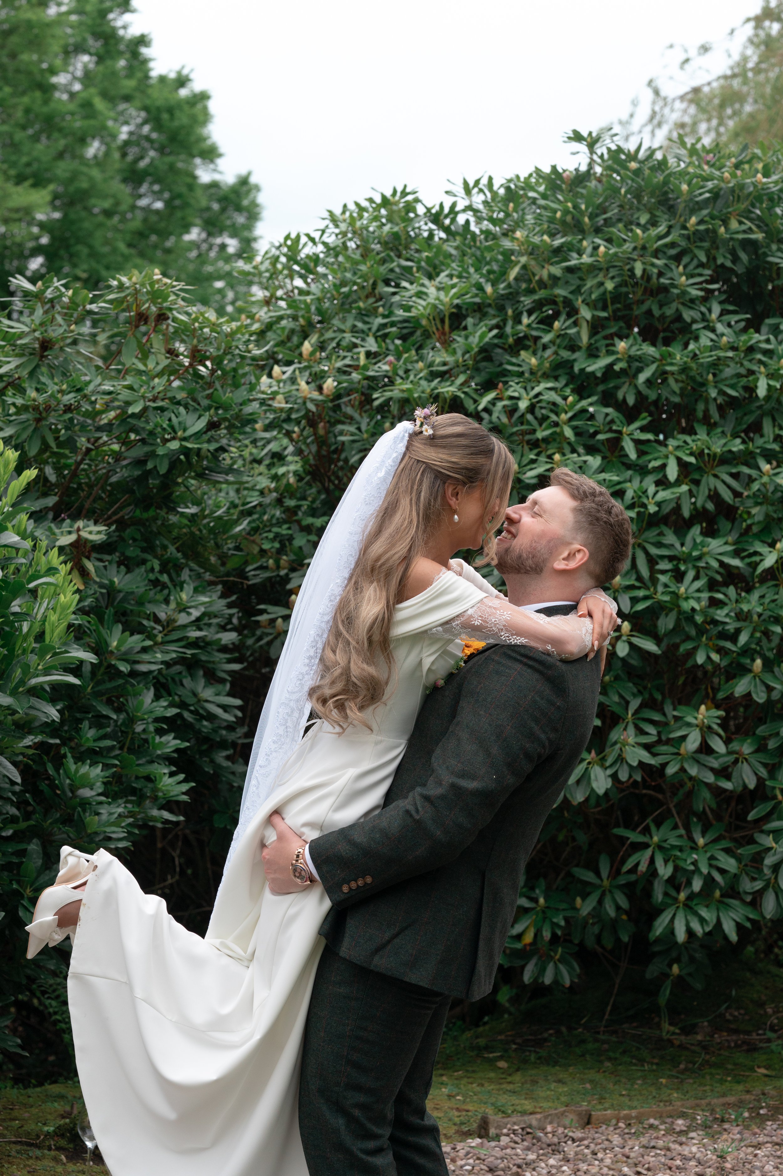 A couple in wedding attire holding each other outdoors surrounded by green bushes and trees, with the woman wearing a white wedding dress and veil, and the man wearing a dark suit.