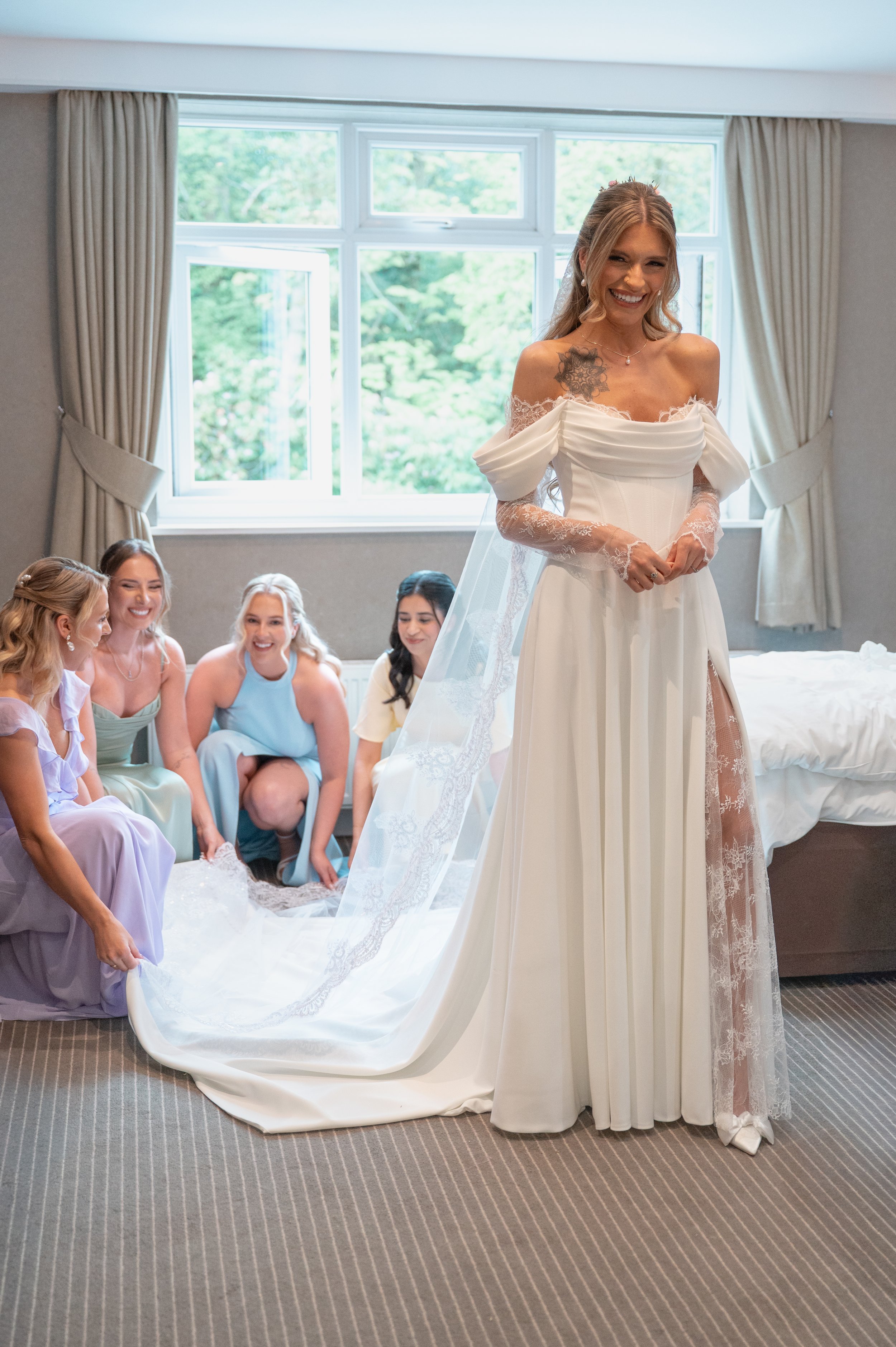 A bride in a wedding dress smiling as she stands in a room with windows, while her friends in colorful dresses sit on the floor and help arrange her train.