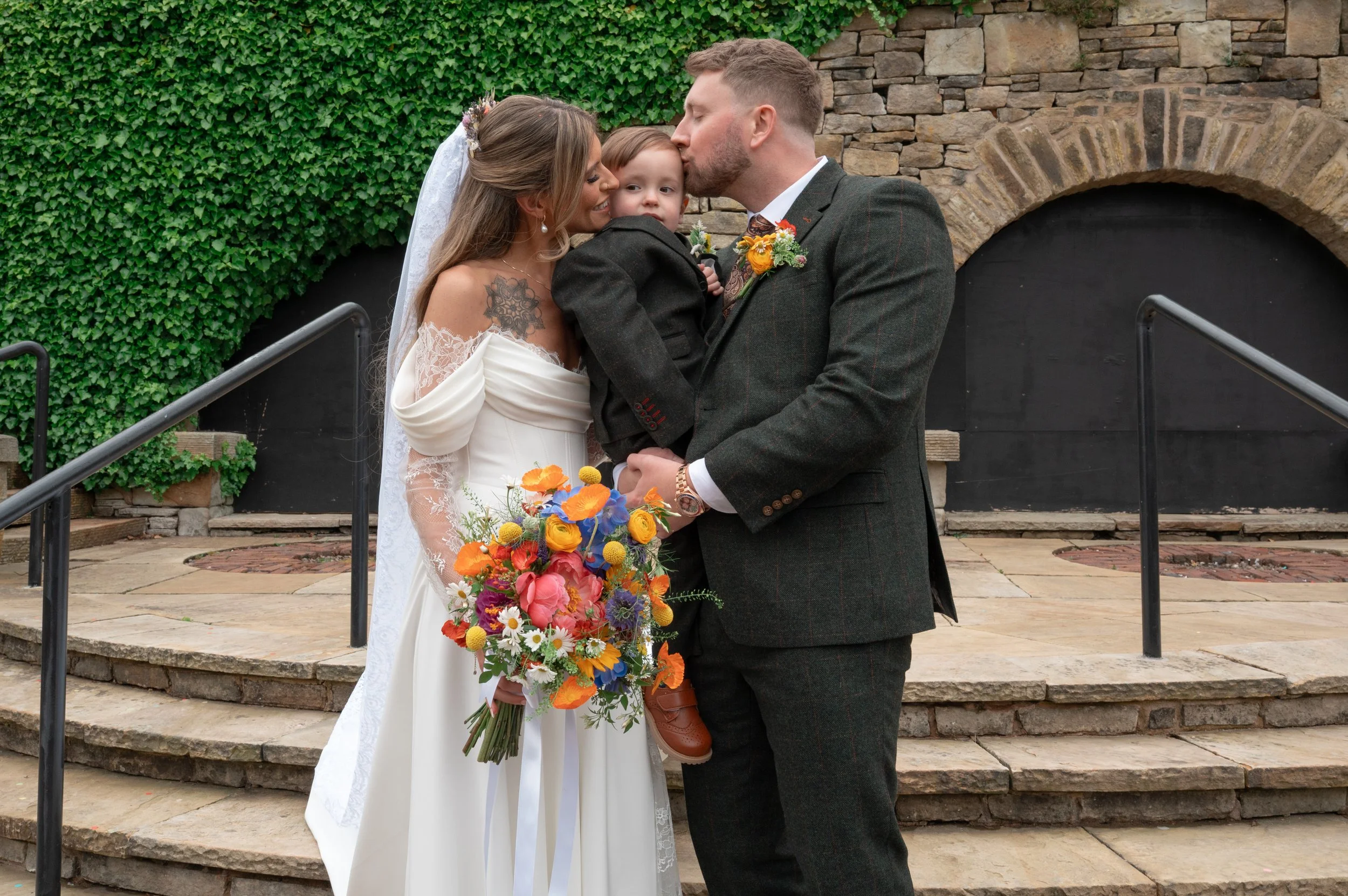A newlywed couple sharing a kiss on their wedding day, with a young boy between them, holding a colorful bouquet, in front of a stone wall with ivy.