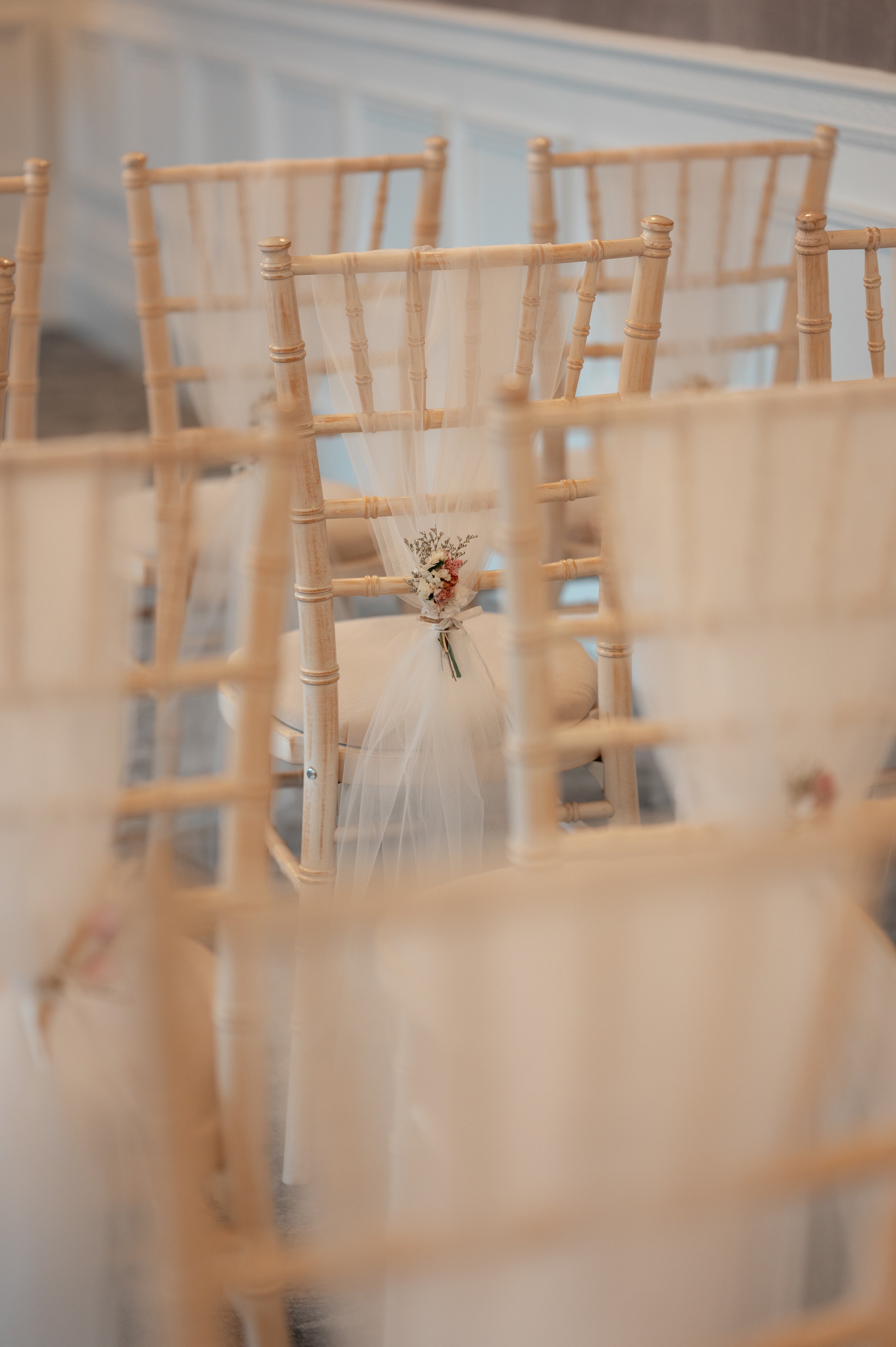 Empty beige chairs in a row at a formal event, with a small floral arrangement on one chair.