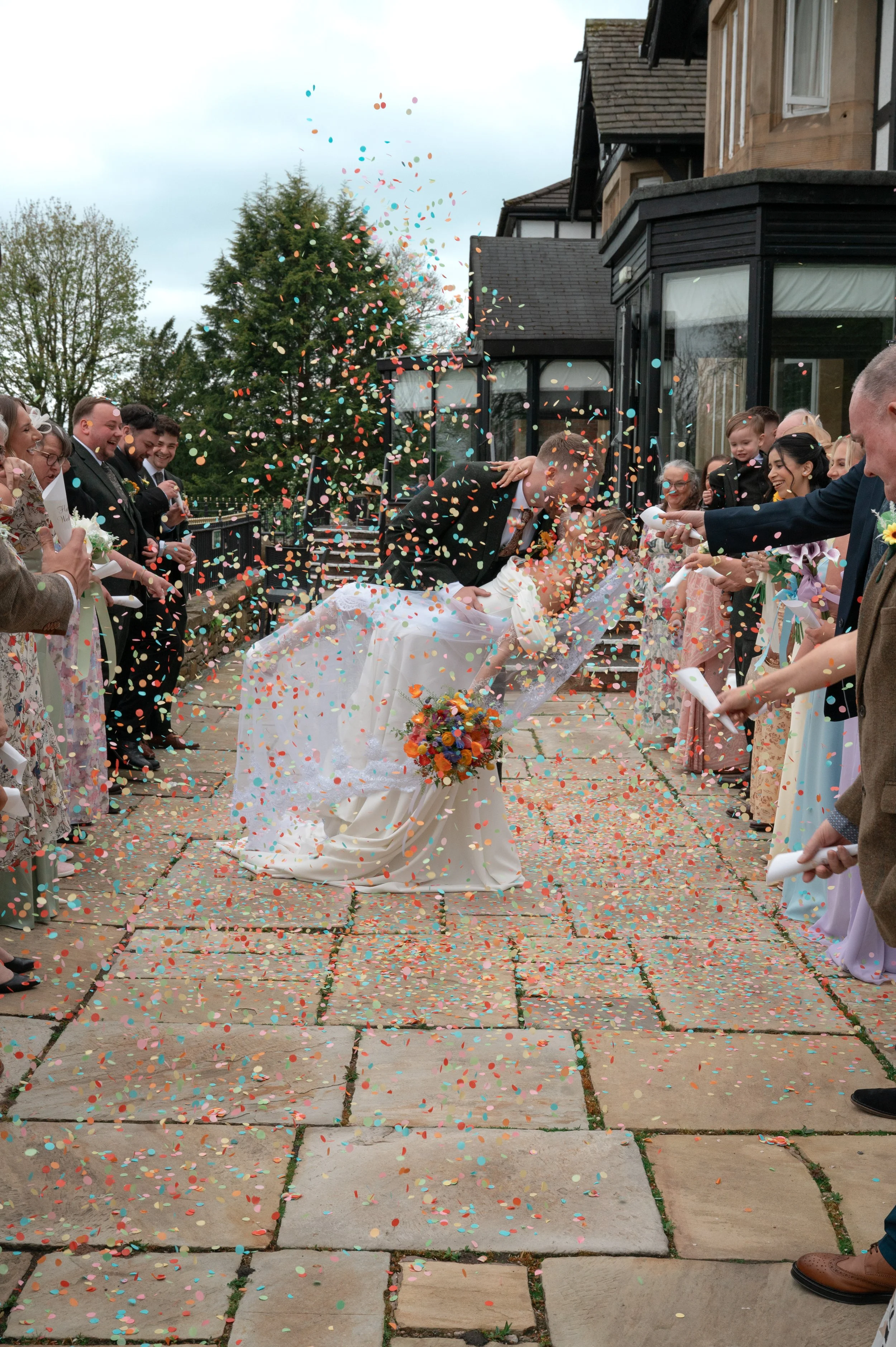 A bride and groom kiss on a brick patio surrounded by guests throwing colorful confetti, celebrating their wedding.