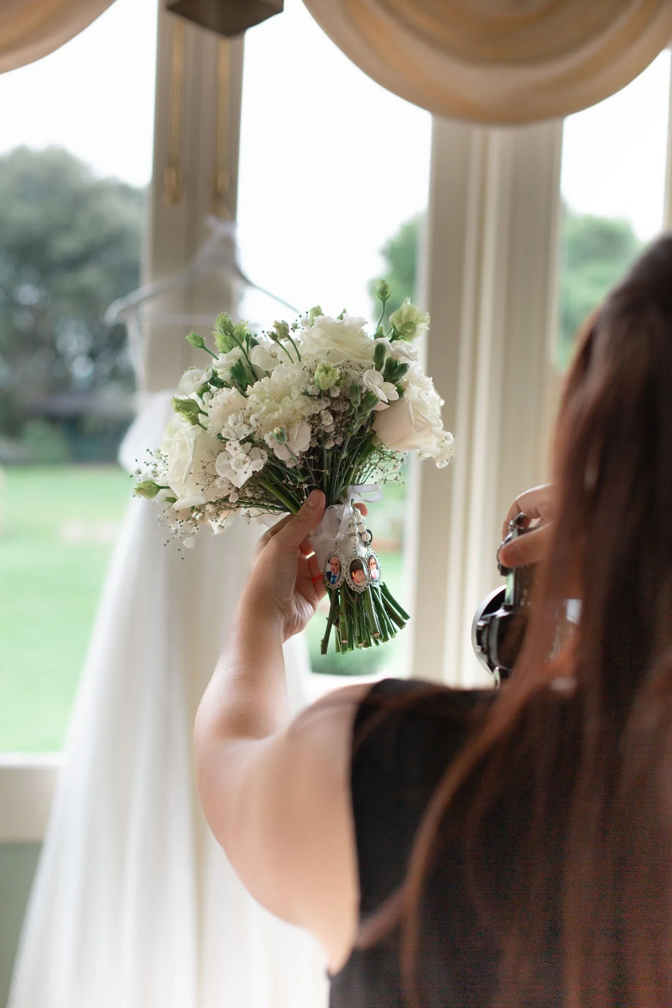 A woman holding a bouquet of white flowers during the daytime near a window with a blurred dress hanging in the background.