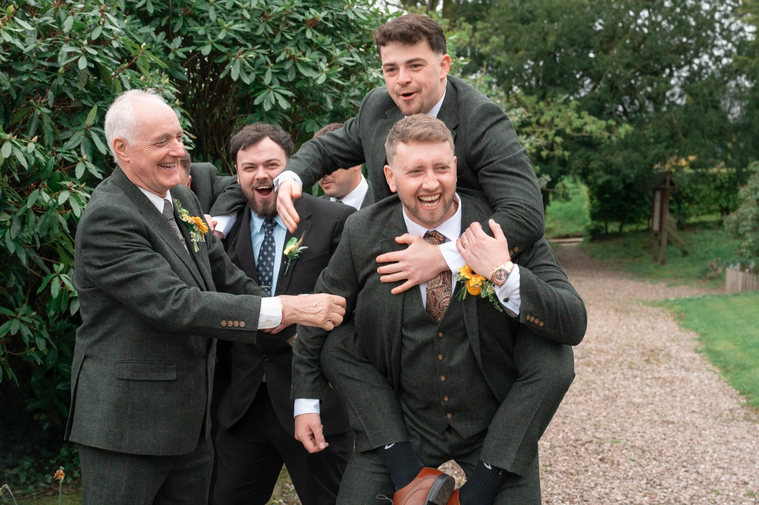 A group of men in suits having fun outdoors, with one man sitting on another's shoulders, all smiling and laughing.