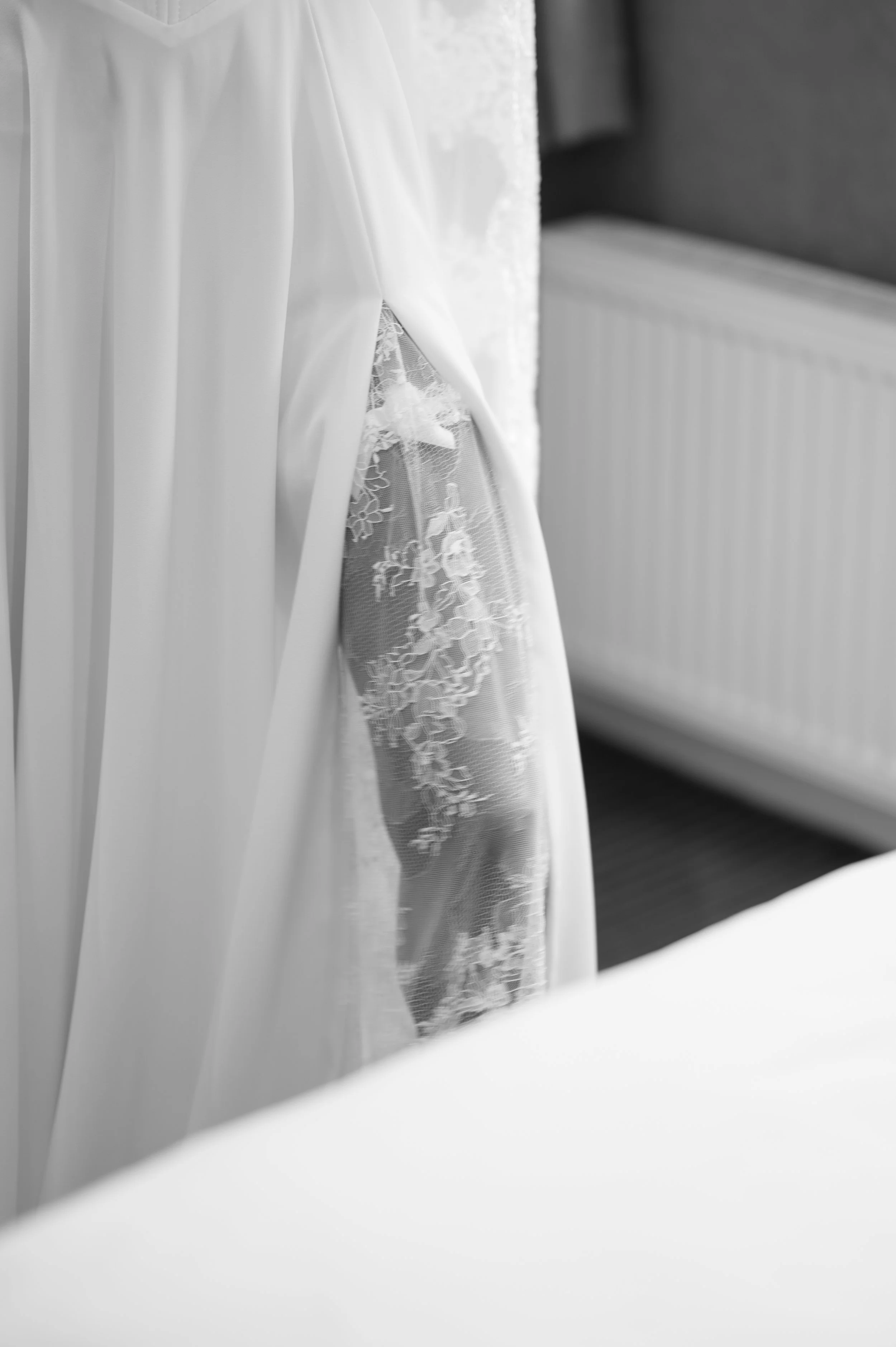 Close-up of a wedding dress hanging, with lace and satin details, seen through a mirror in a room with a radiator.