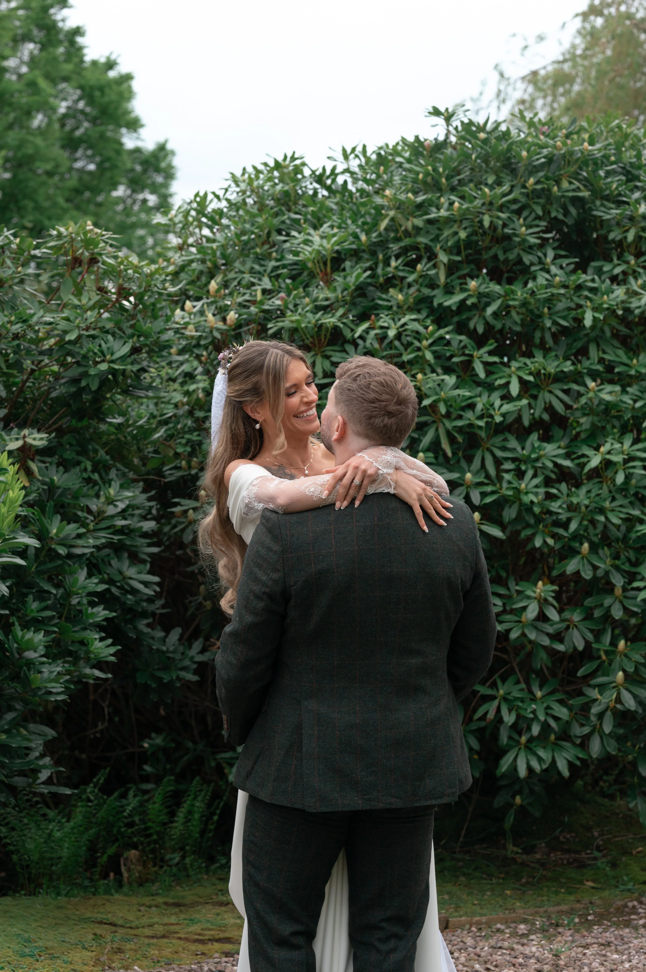 A woman in a wedding dress and a man in a suit embracing outdoors, surrounded by greenery.