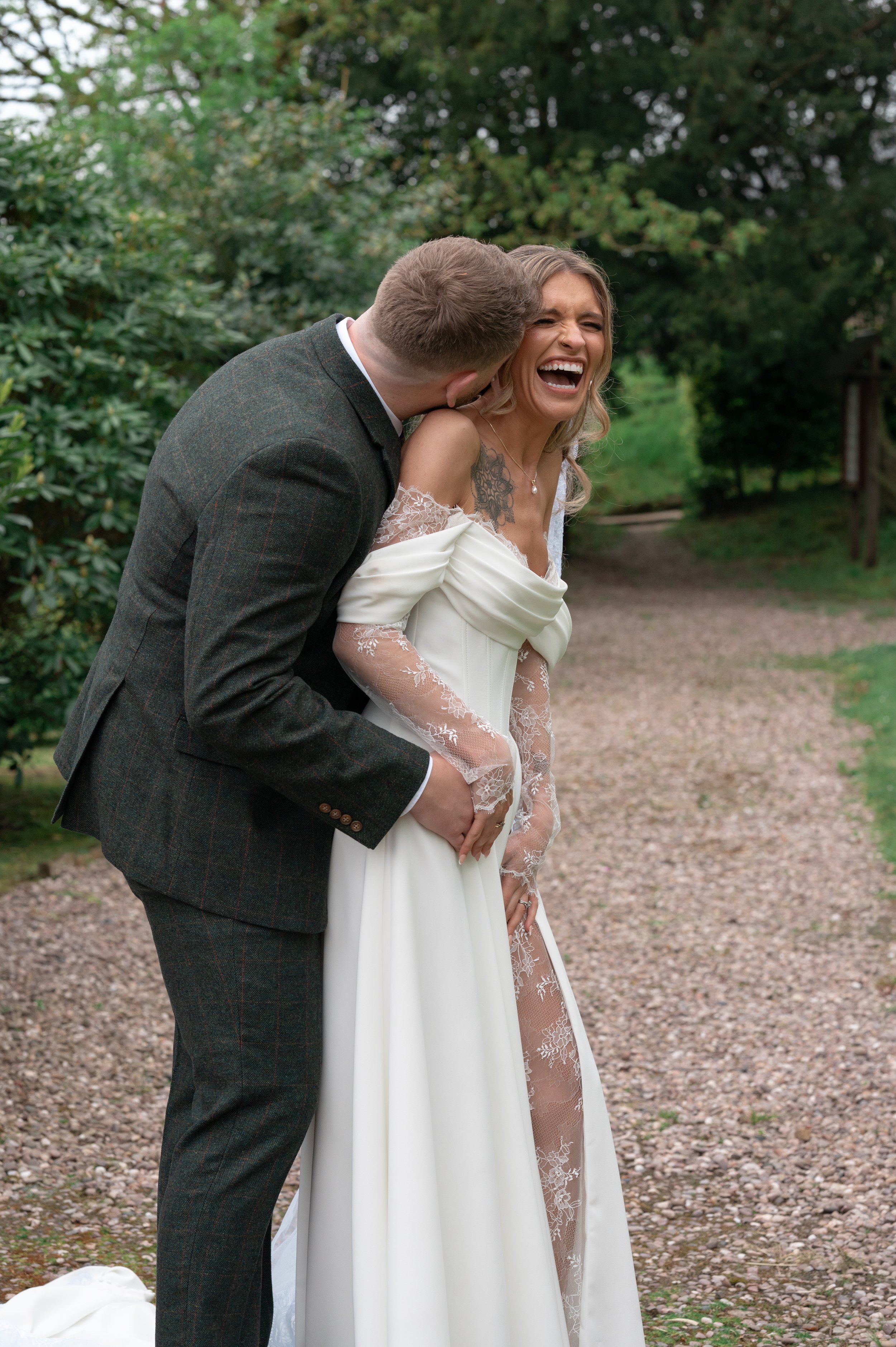 A bride and groom sharing a funny moment outdoors on a gravel path surrounded by greenery. The groom leans in towards the bride's face, and the bride is laughing with her mouth open, showing her teeth. The bride is wearing a white dress with lace det