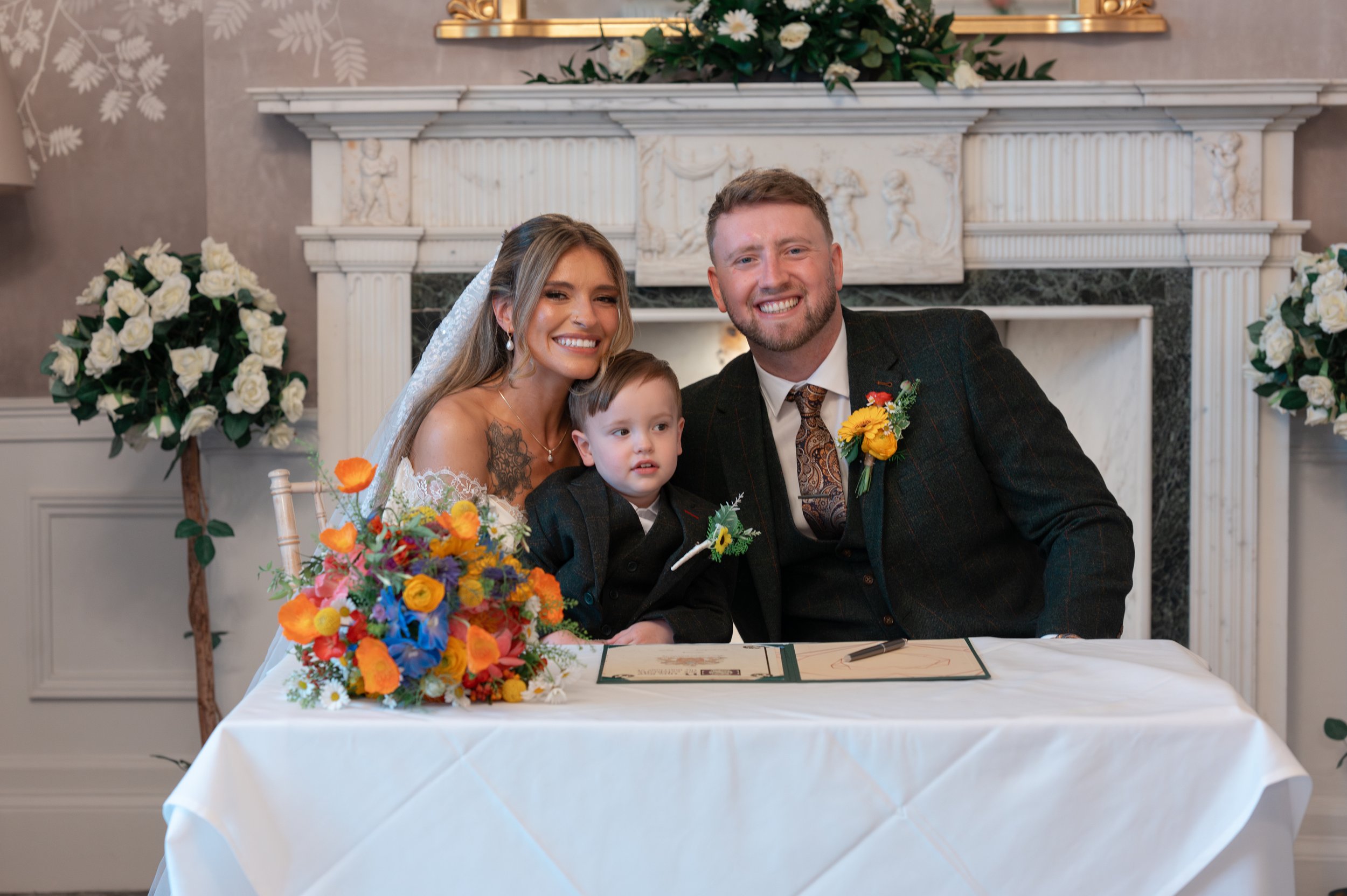 A smiling couple, a woman and a man, with a young boy, sitting at a table with a colorful floral arrangement, signing a document in front of a decorative fireplace with floral arrangements on each side.