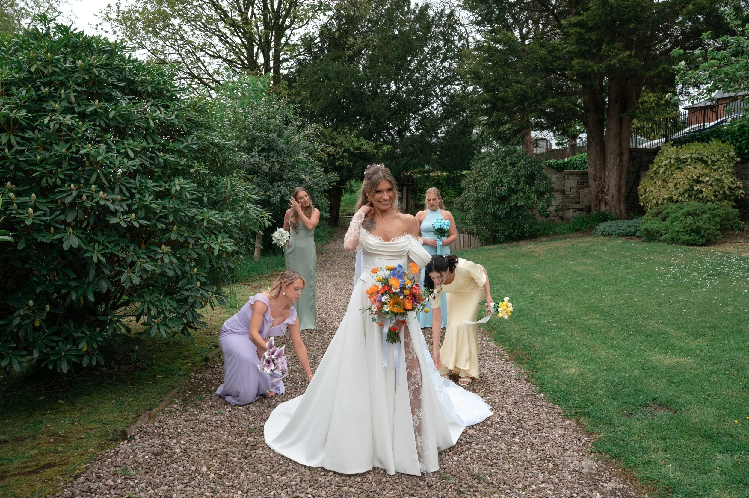 A bride in a white wedding dress holding a colorful bouquet, walking on a gravel path outdoors, with bridesmaids in pastel dresses around her, surrounded by green trees and bushes.
