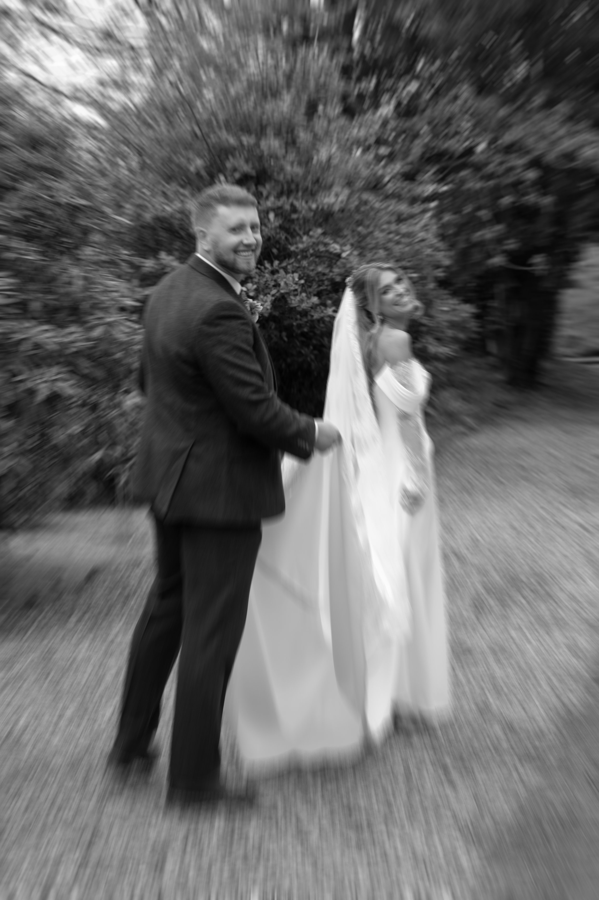 A black and white photo of a smiling bride and groom holding hands, walking outdoors with a blurred background of trees and bushes, with a radial blur effect.