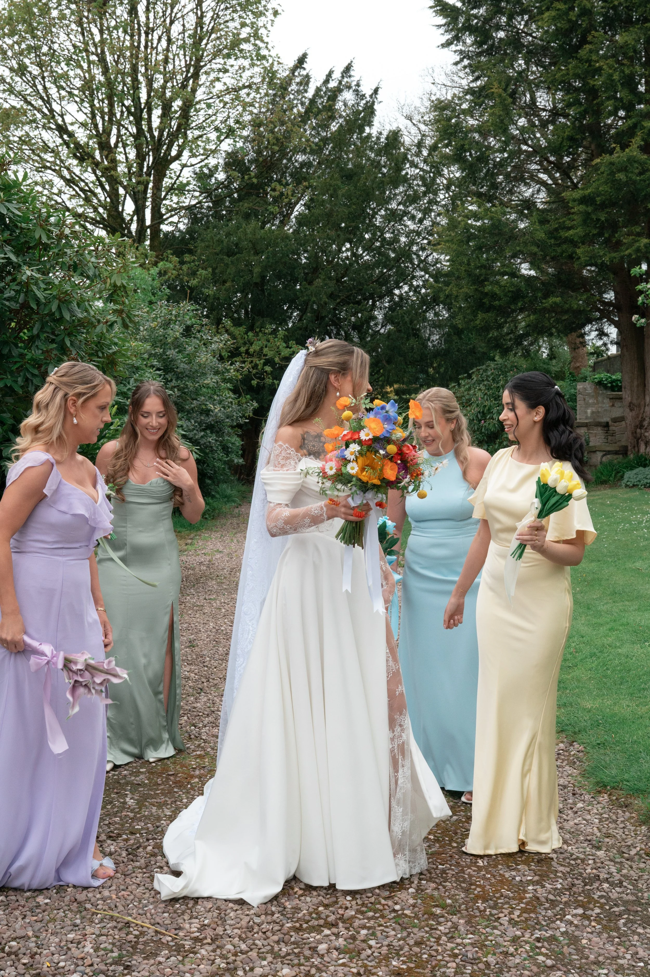 A bride in a white lace wedding gown holding a colorful bouquet standing with five women in pastel-colored dresses, holding flowers, outdoors with trees and cloudy sky in the background.
