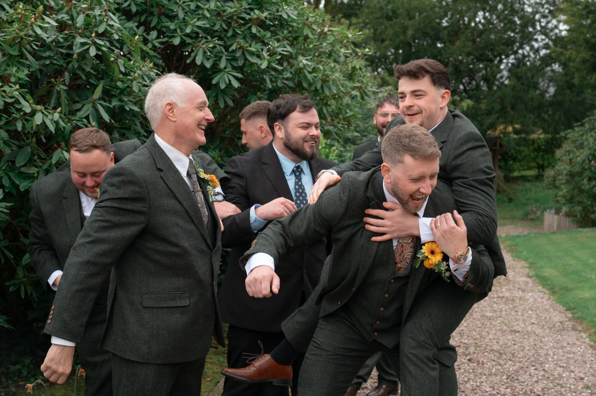 A group of men in suits playing a game that resembles a traditional British game called planking, with one man laying horizontally on the back of another, while others support and laugh around them outdoors.