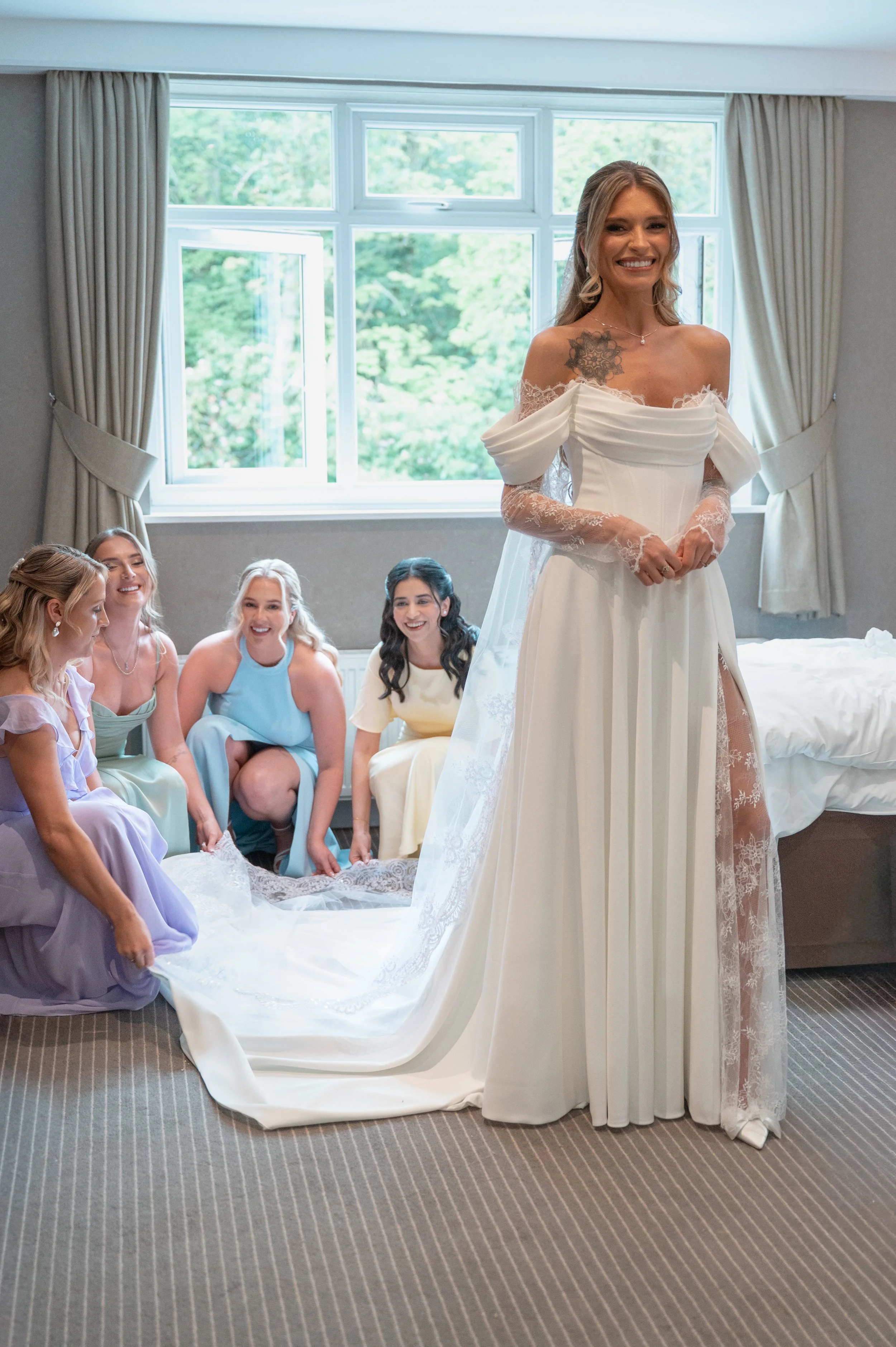 A bride in a white wedding gown standing in front of a group of women seated on the floor, all inside a room with a large window and cream-colored curtains, during a bridal preparation or celebration.