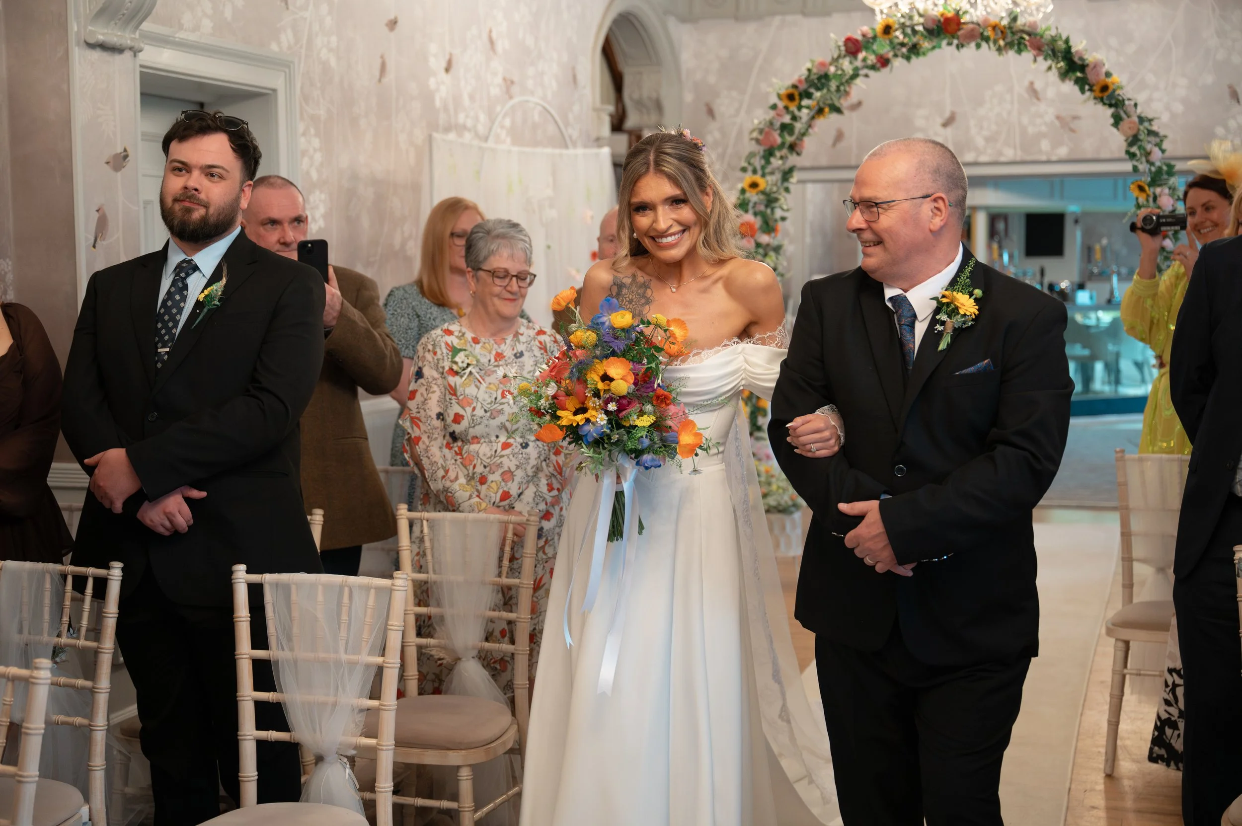 A bride smiling and holding a colorful bouquet of flowers, standing between two men in suits during a wedding ceremony, with a floral arch and seated guests in the background.