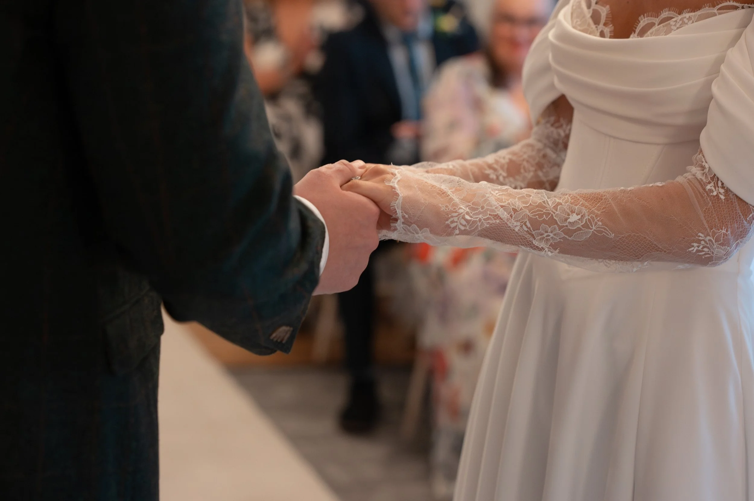 Close-up of a couple holding hands during a wedding ceremony, with the bride wearing a lace wedding dress and the groom in a dark suit. Blurred guests are visible in the background.