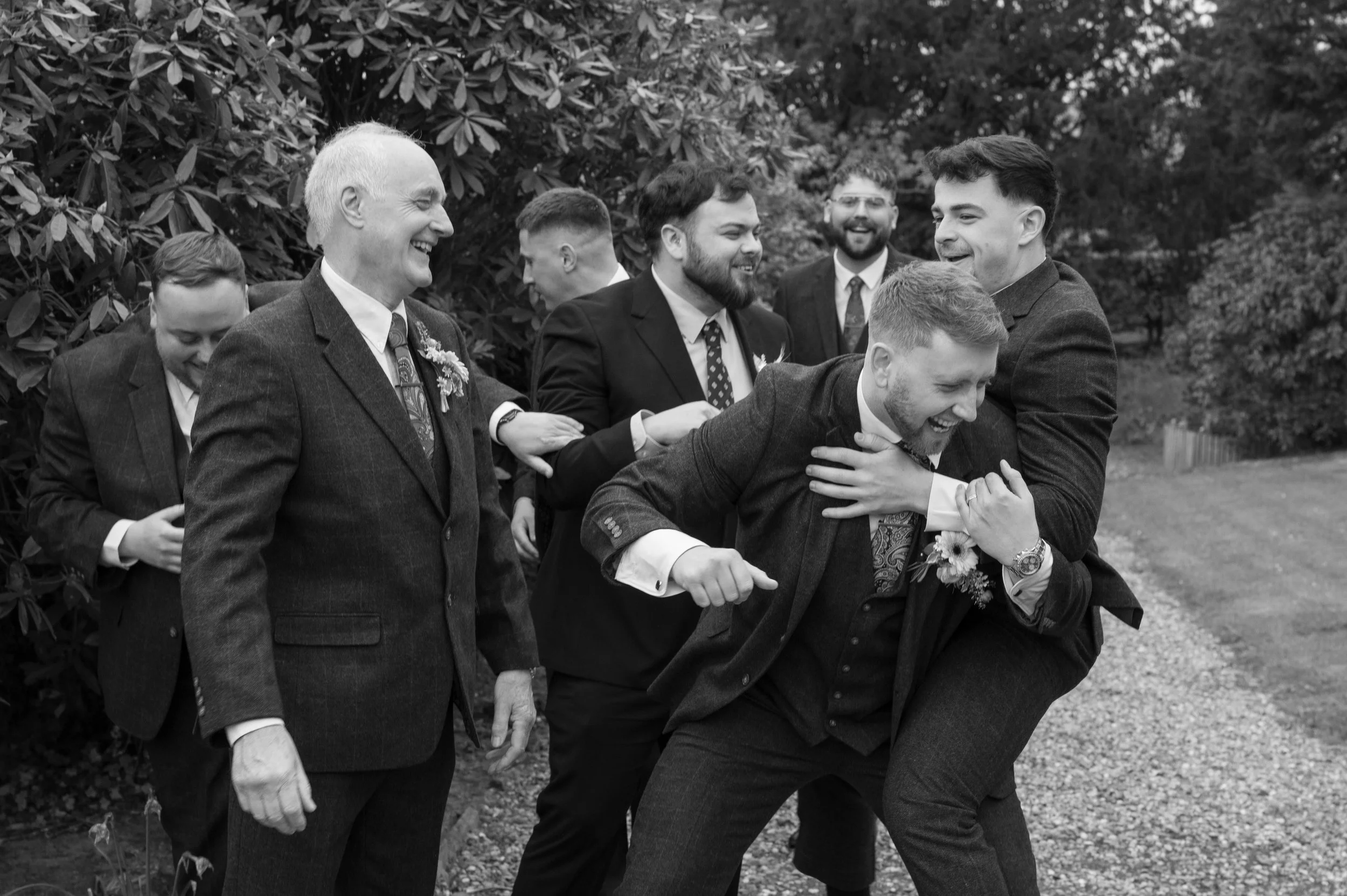 A group of men in suits laughing and playing together outdoors, possibly at a wedding or celebration, with trees in the background.