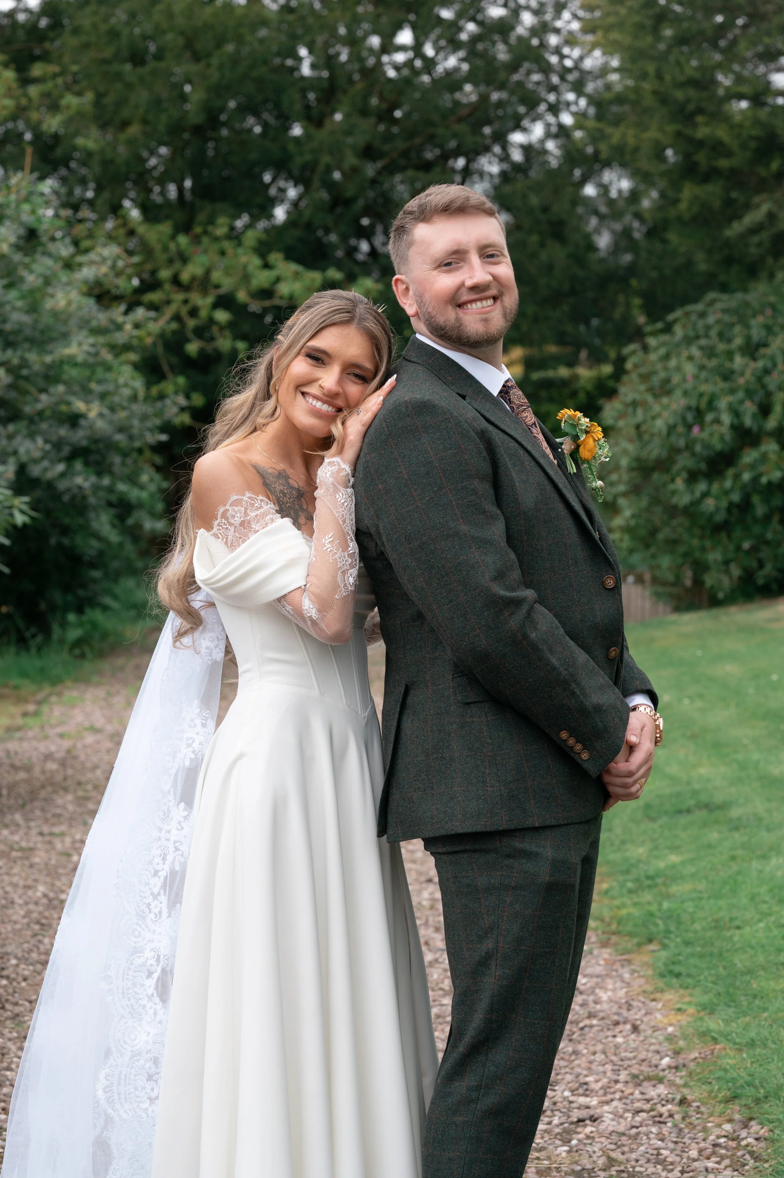 A bride and groom posing outdoors on a gravel path, with trees and greenery in the background. The bride is wearing a white dress with lace details and the groom is dressed in a dark checked suit with a boutonniere.