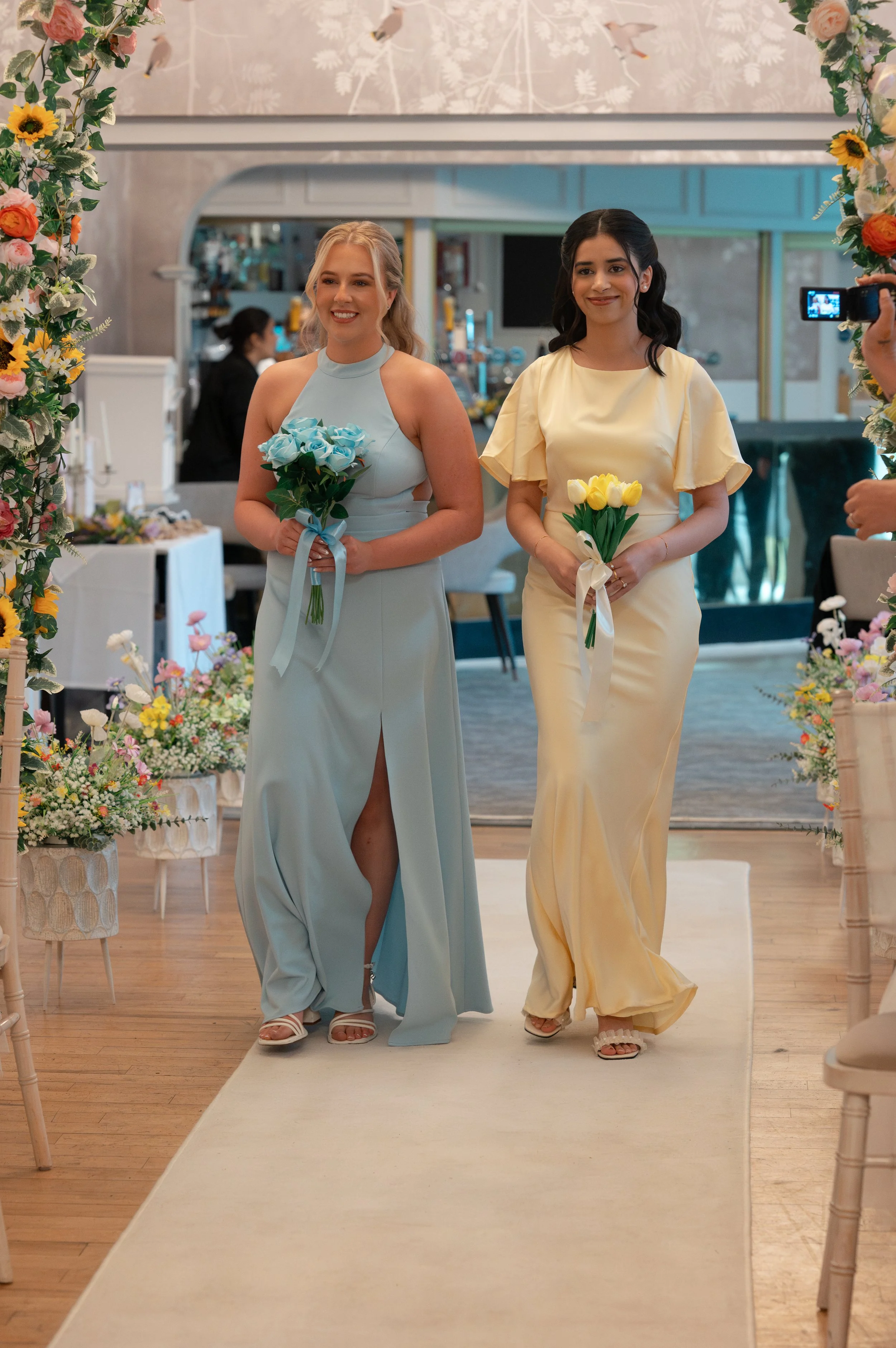 Two women in formal dresses walking down an aisle decorated with flowers, holding bouquets, at a wedding or similar event.