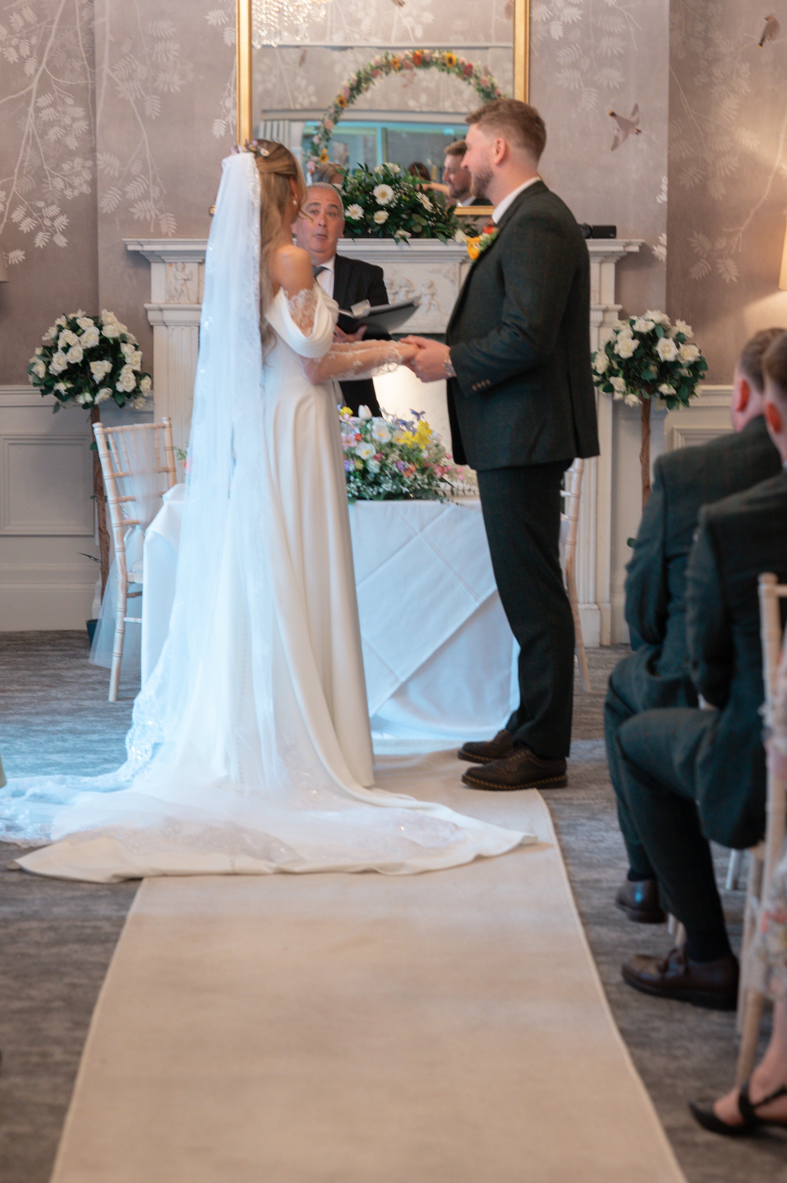 A wedding ceremony with a bride and groom holding hands, facing an officiant. The bride is wearing a white dress with lace sleeves and a veil, while the groom is in a dark suit. Flowers and a mirror decorate the background, and guests are seated on t