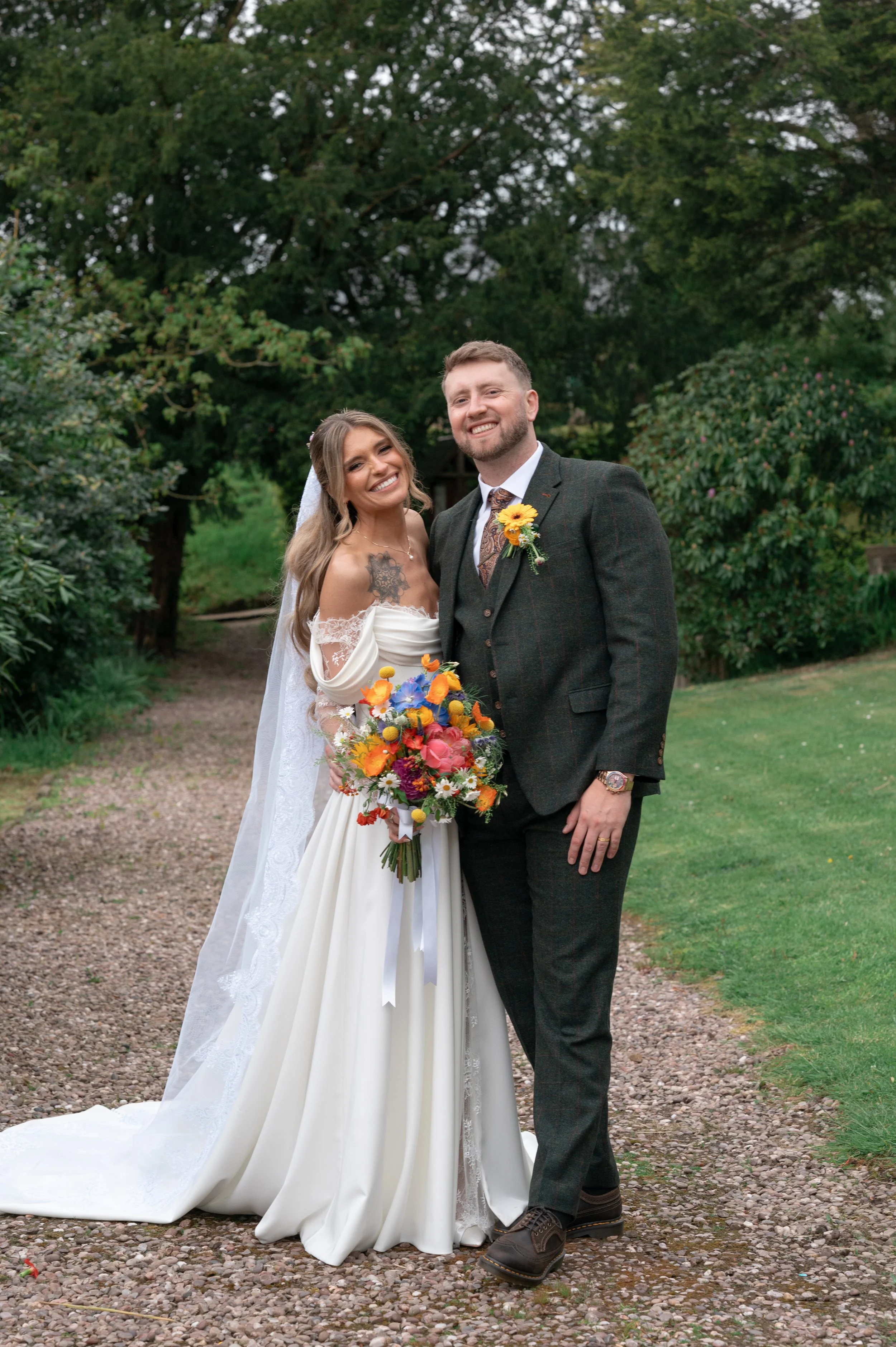A bride and groom standing outdoors on a gravel path, smiling for the camera, with trees in the background. The bride is wearing a white wedding dress with lace details and holding a colorful bouquet of flowers. The groom is dressed in a dark suit wi