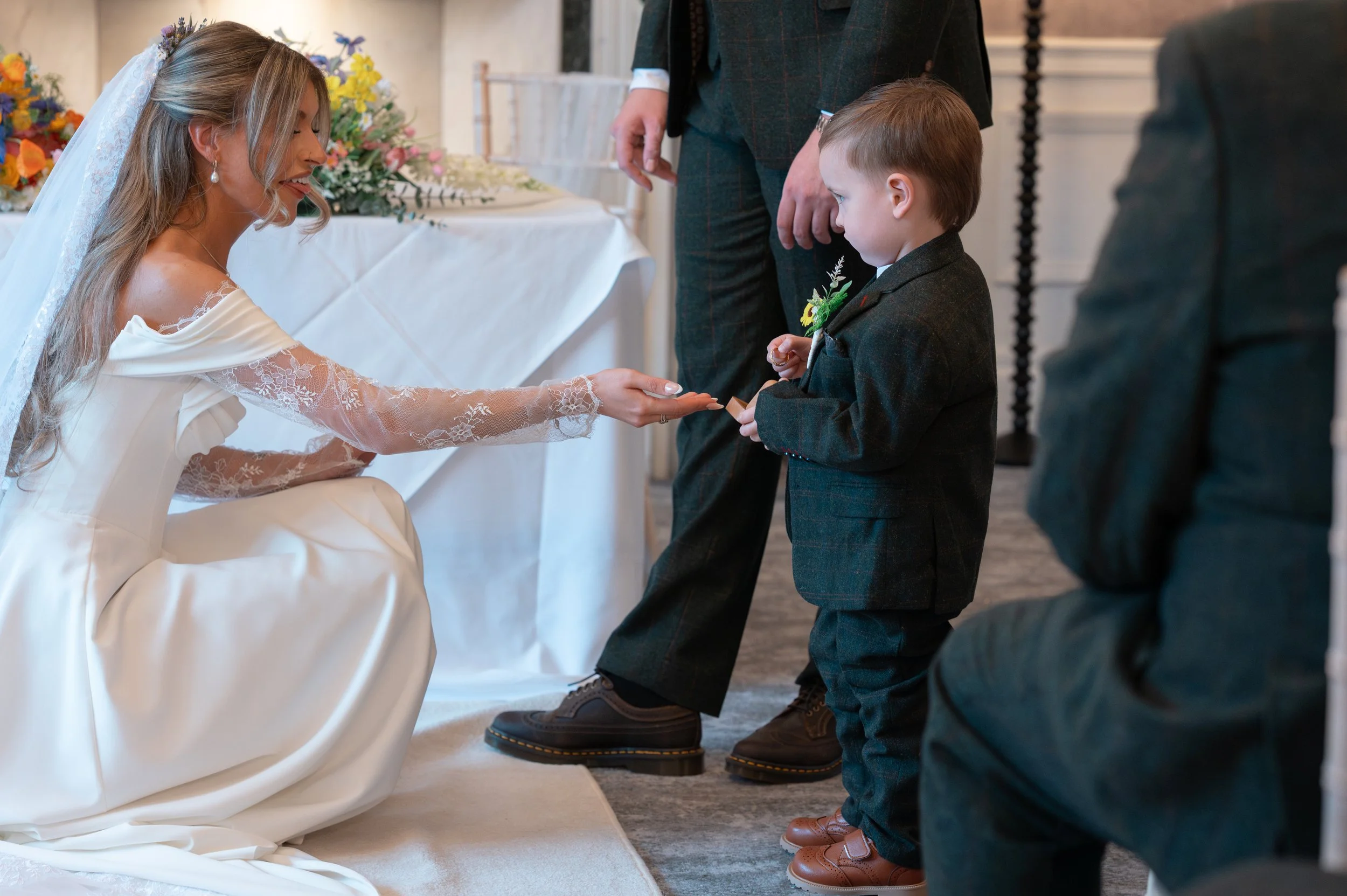 A bride in a white wedding dress kneels on the floor, holding hands with a young boy dressed in a suit, during a wedding ceremony. The bride has long, wavy hair with a veil and pearl earrings. The boy has brown hair and a boutonniere on his suit. The