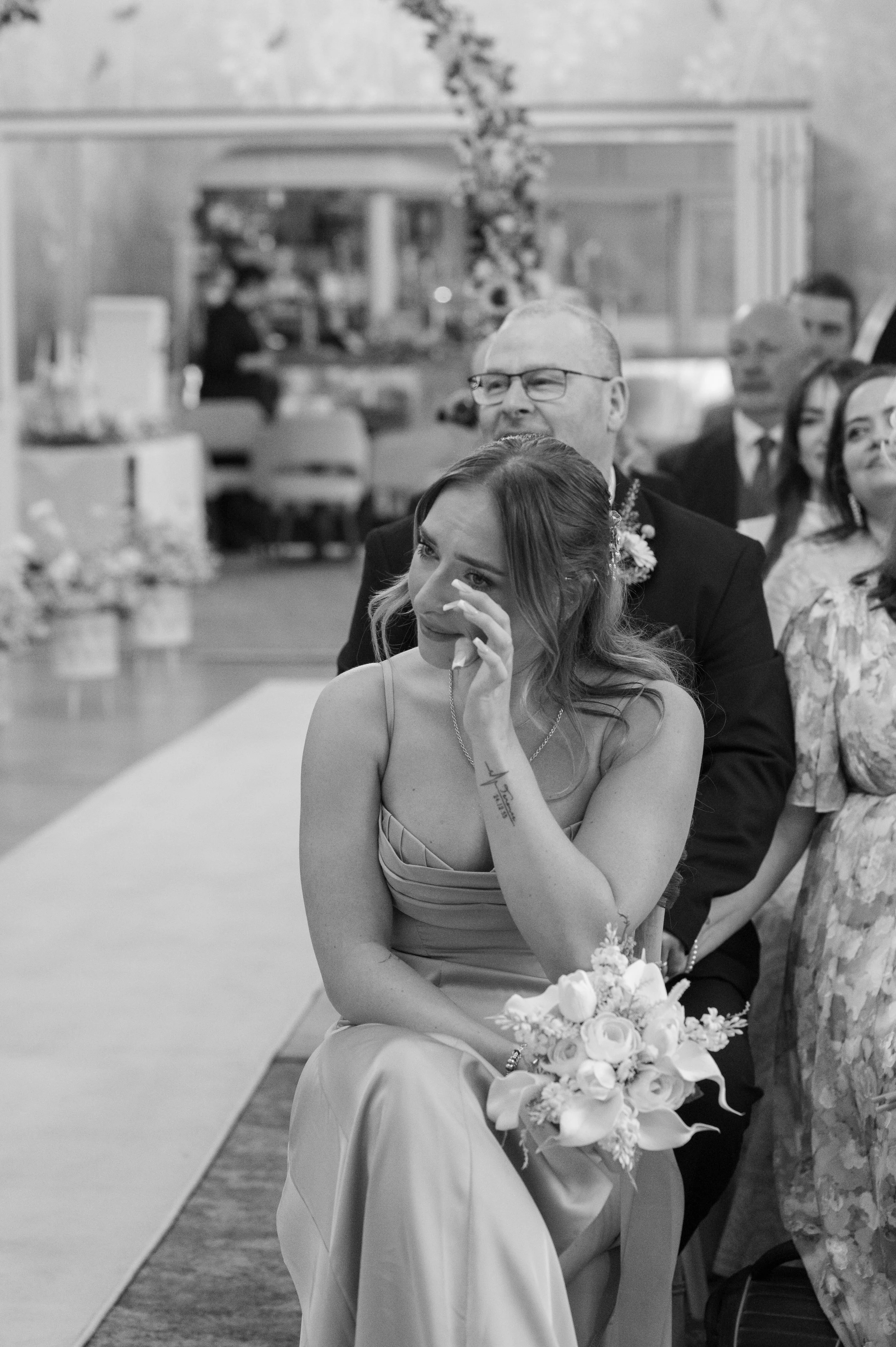 A woman in a dress holding a bouquet of white flowers sitting in a row of guests listening at a wedding ceremony, with a man with glasses and a suit sitting behind her, and other guests visible in the background.