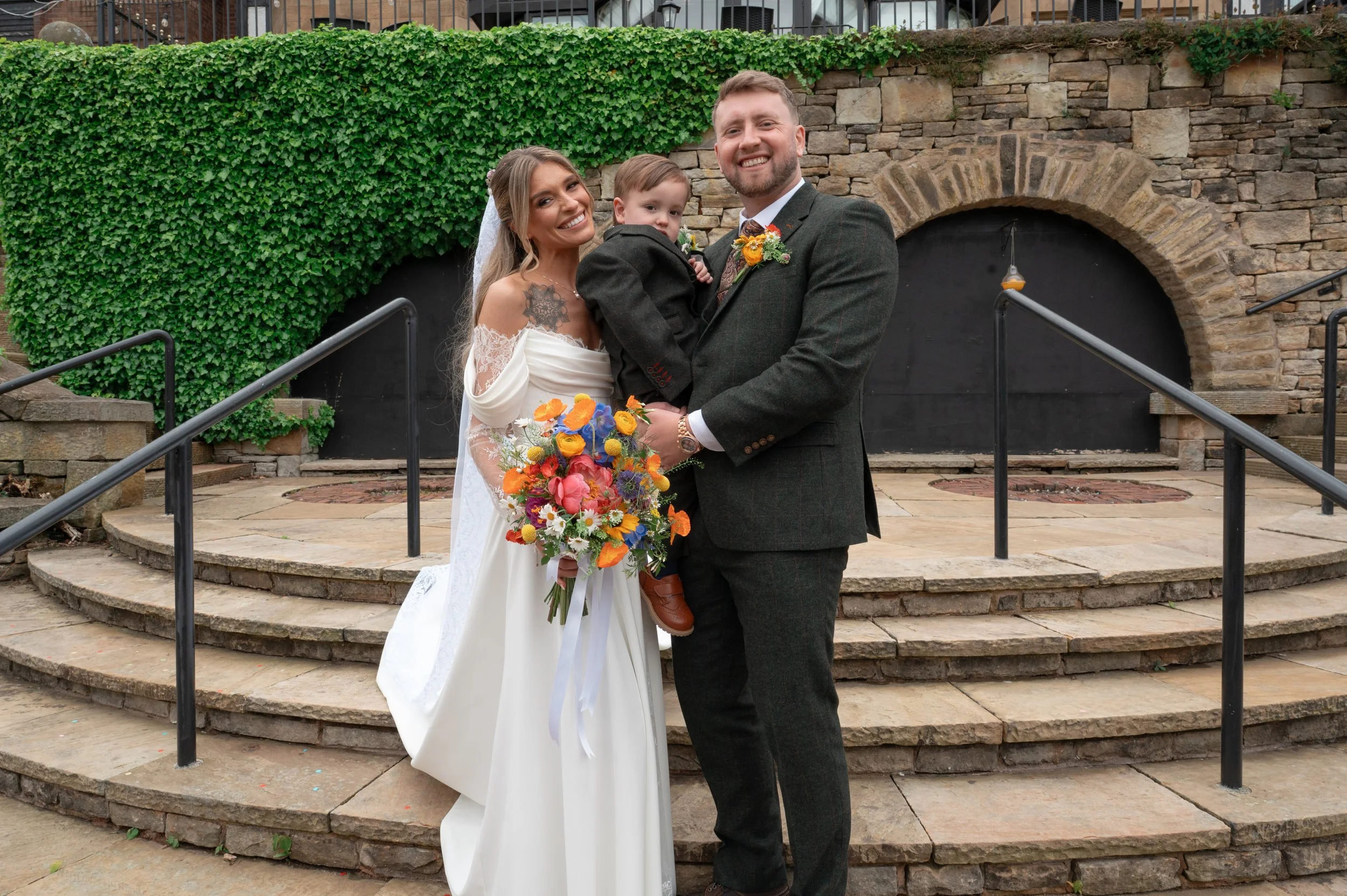A wedding couple stands on stone steps outside, holding a young boy between them. The bride is in a white off-shoulder wedding dress with lace sleeves and a veil, holding a colorful bouquet. The groom is in a dark gray suit with a floral boutonniere.