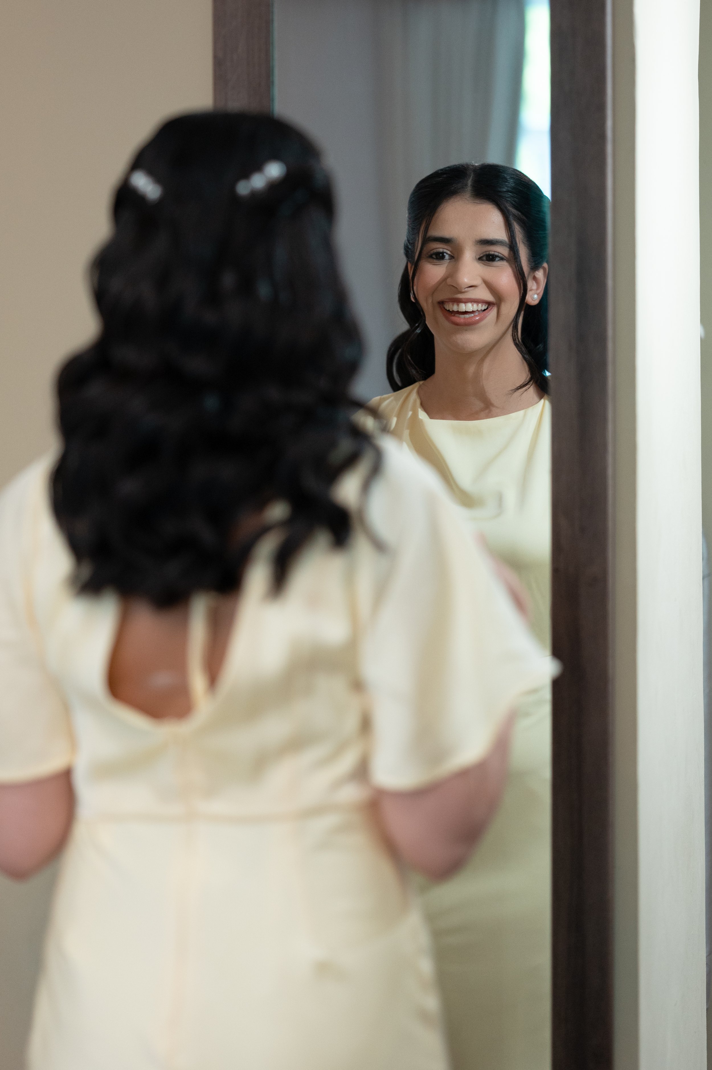 A woman with dark hair styled in loose curls, wearing a light-colored off-shoulder dress, is smiling at herself in a mirror.