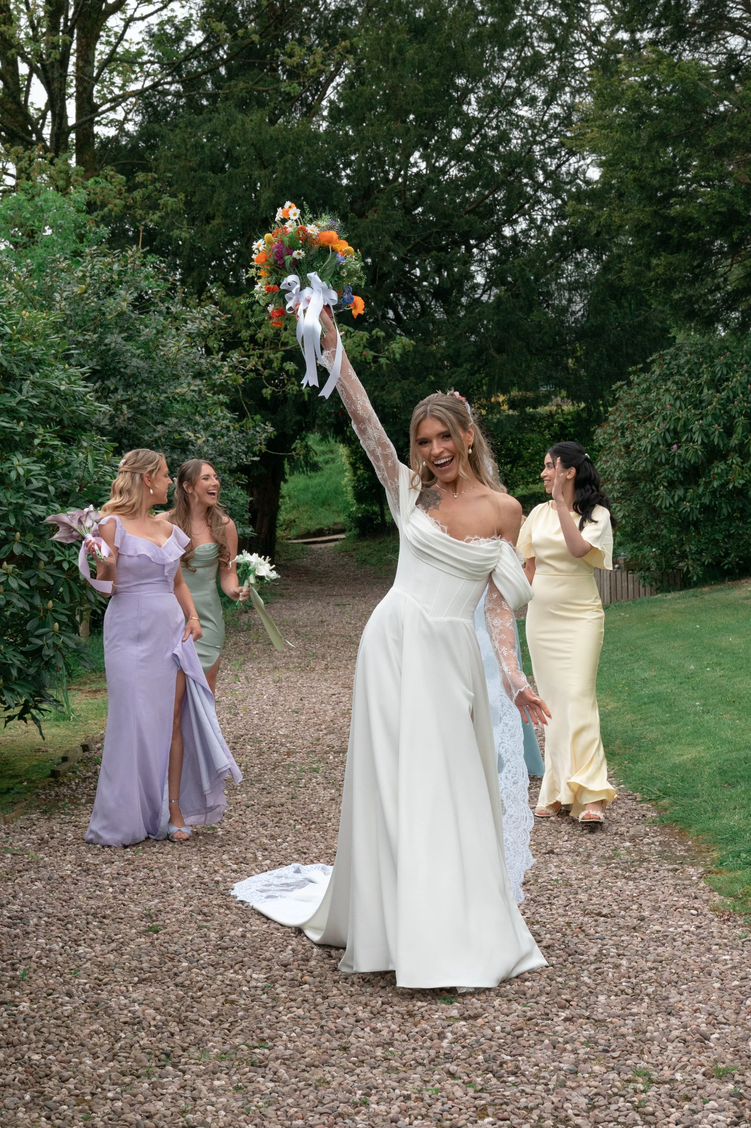 A bride in a white wedding dress holding a bouquet of orange, yellow, and white flowers high above her head, smiling. Four women in pastel-colored dresses are standing behind her outdoors on a gravel path, surrounded by green trees and bushes, celebr