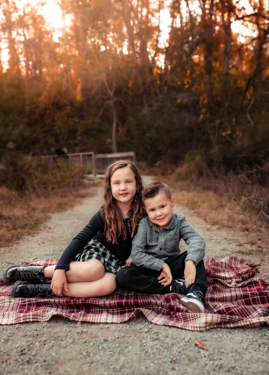 A young girl and boy sitting together on a plaid blanket outdoors during sunset, surrounded by trees.