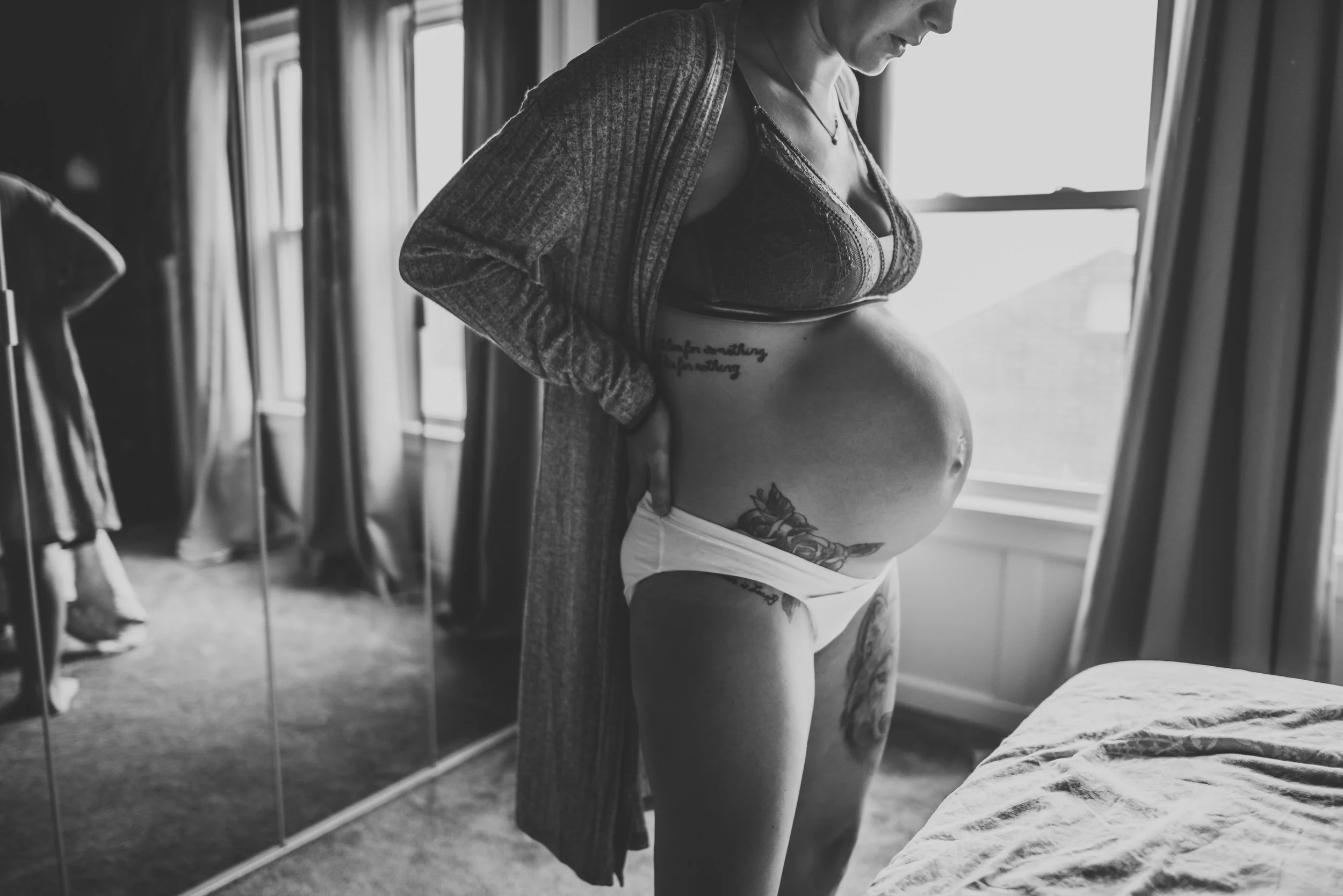 A pregnant woman wearing a bra and underwear, standing by a window and looking down, with tattoos on her belly and thigh, in a bedroom with curtains and a wardrobe.
