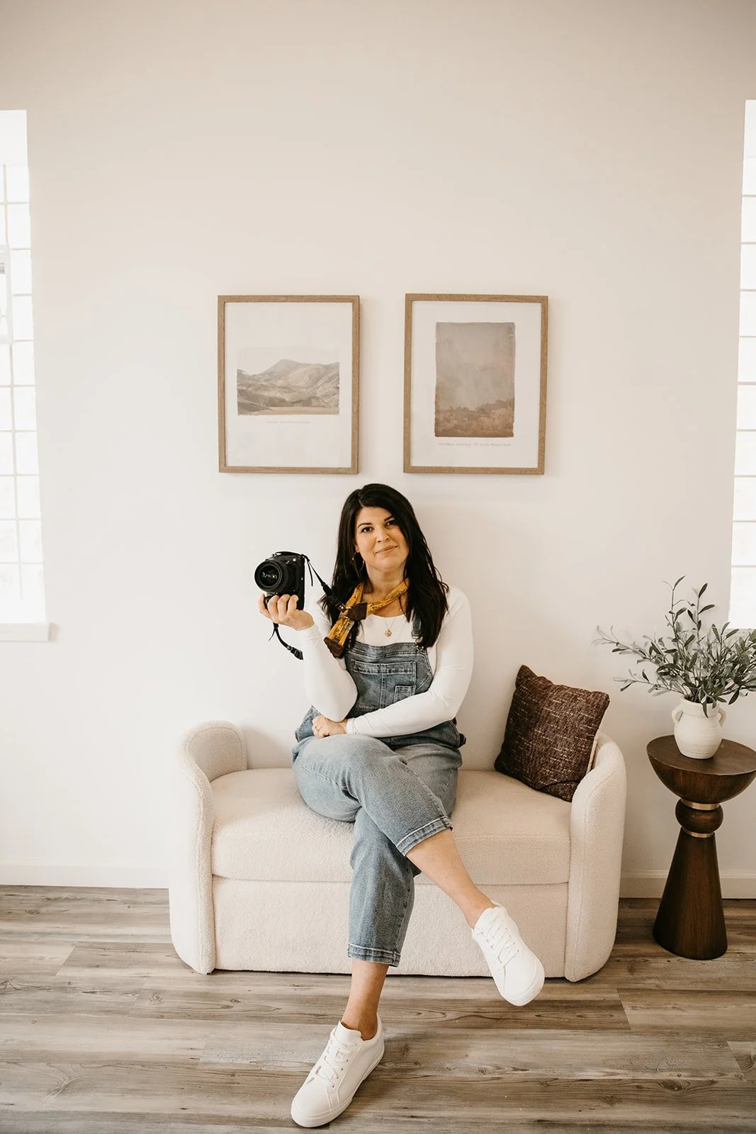 A woman sitting on a cream-colored sofa holding a camera in one hand, wearing a white long-sleeve shirt, gray overalls, and white sneakers. There are two framed pictures hanging on the white wall behind her, a dark pillow on the sofa, and a small table with a potted plant to her right.