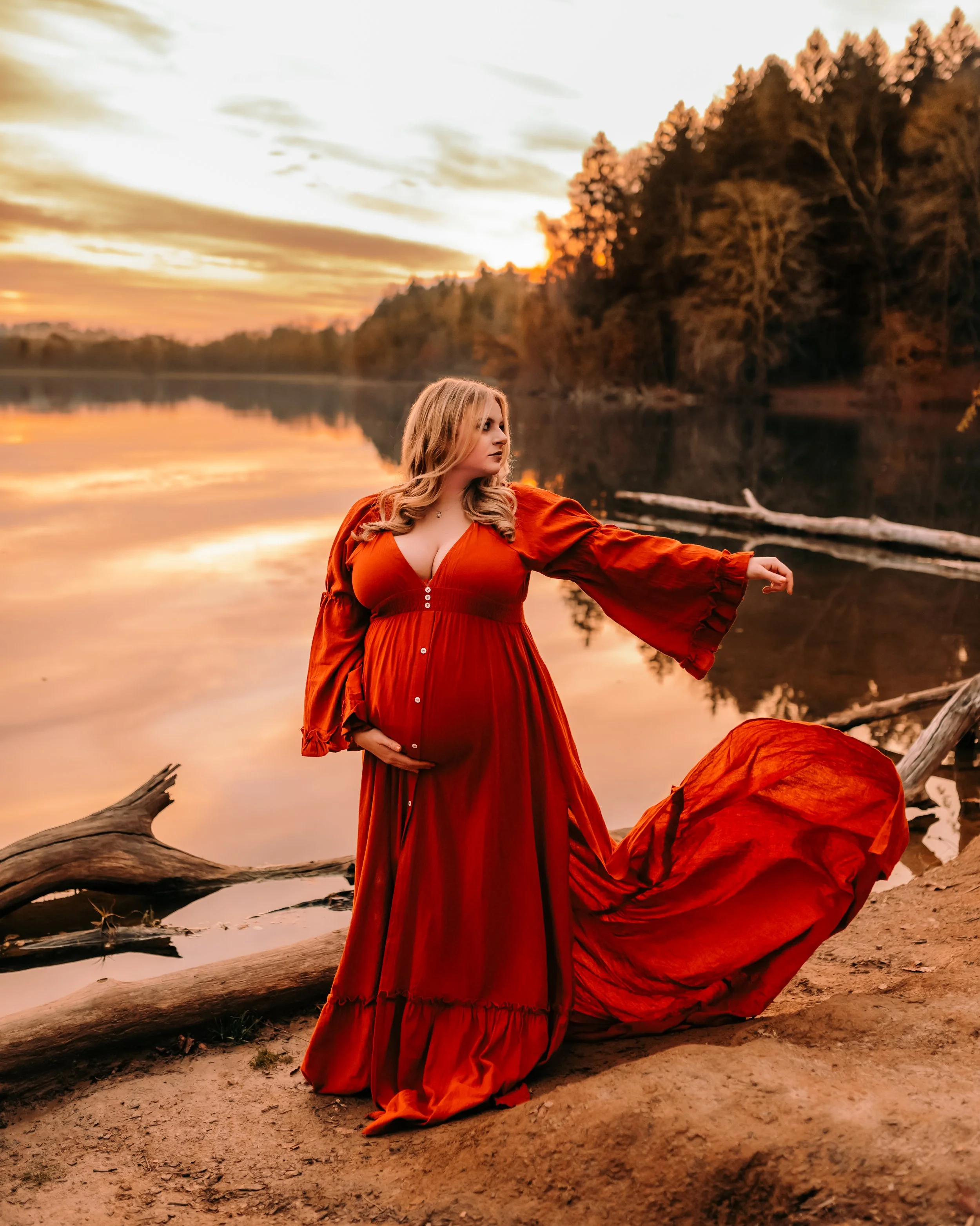 A woman in a red dress standing on a sandy riverbank during sunset, with trees and driftwood in the background.