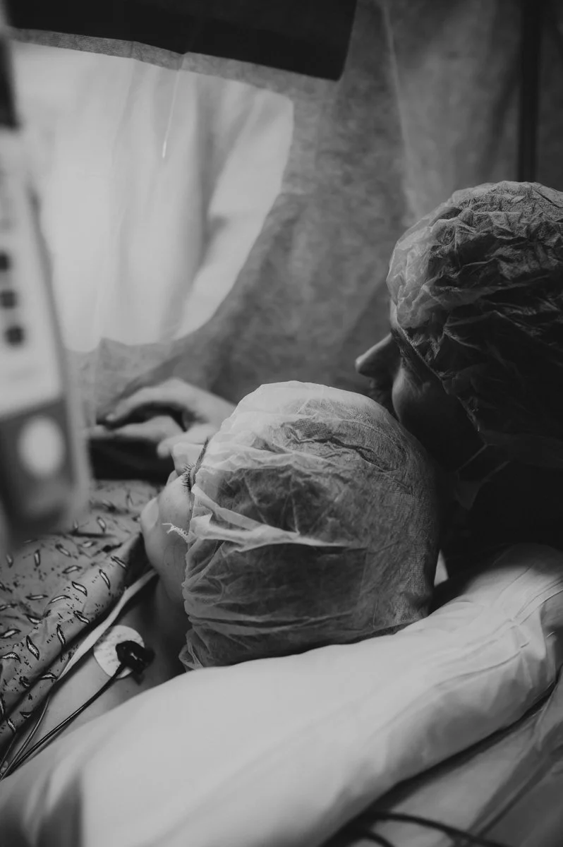 A person in a hospital bed wearing a medical cap, with a healthcare worker in protective gear beside them in a hospital room.