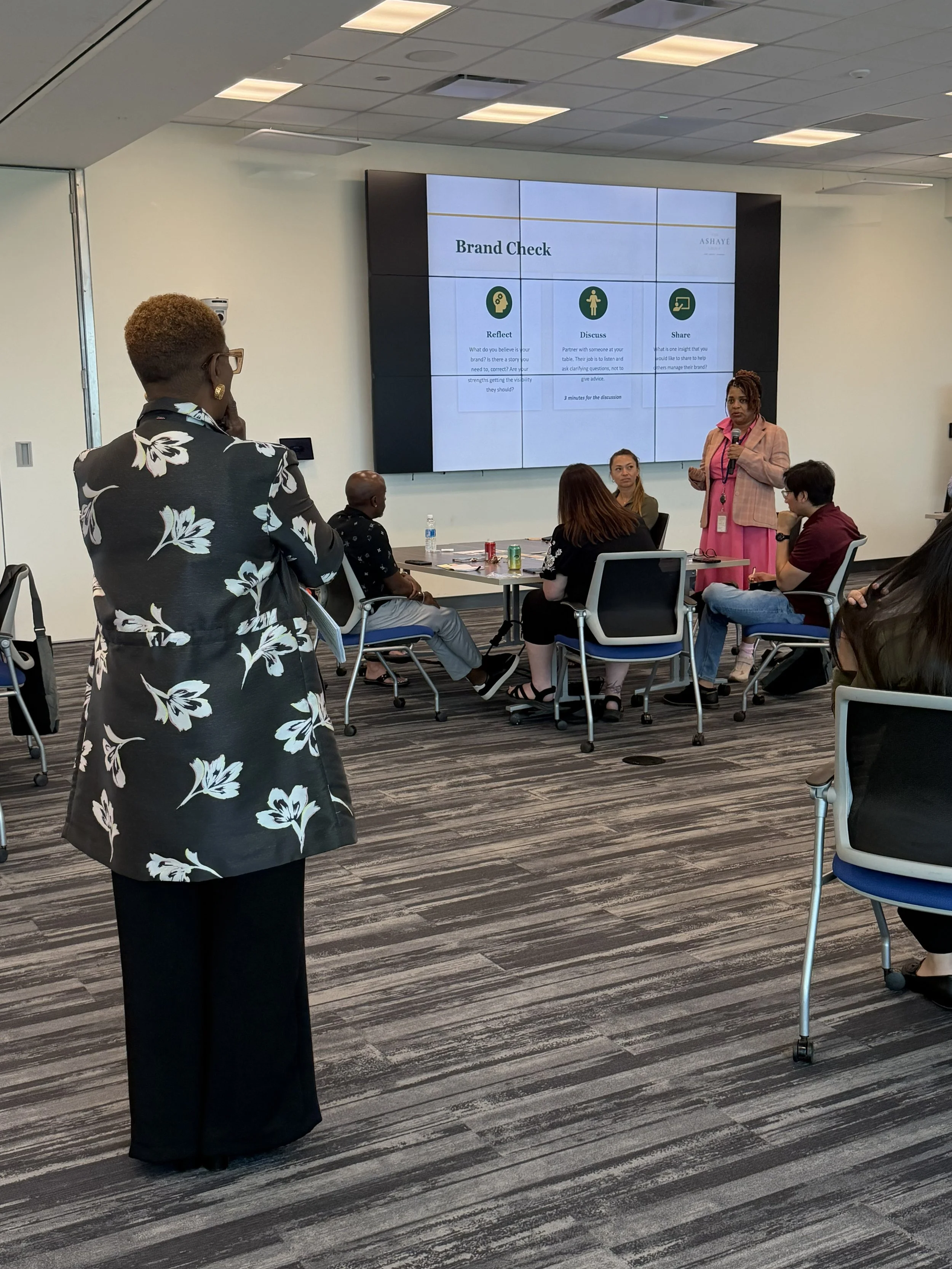 A woman stands in front of a screen displaying a presentation titled 'Brand Check' with instructions to reflect, discuss, and share. Other people are seated at tables, listening.