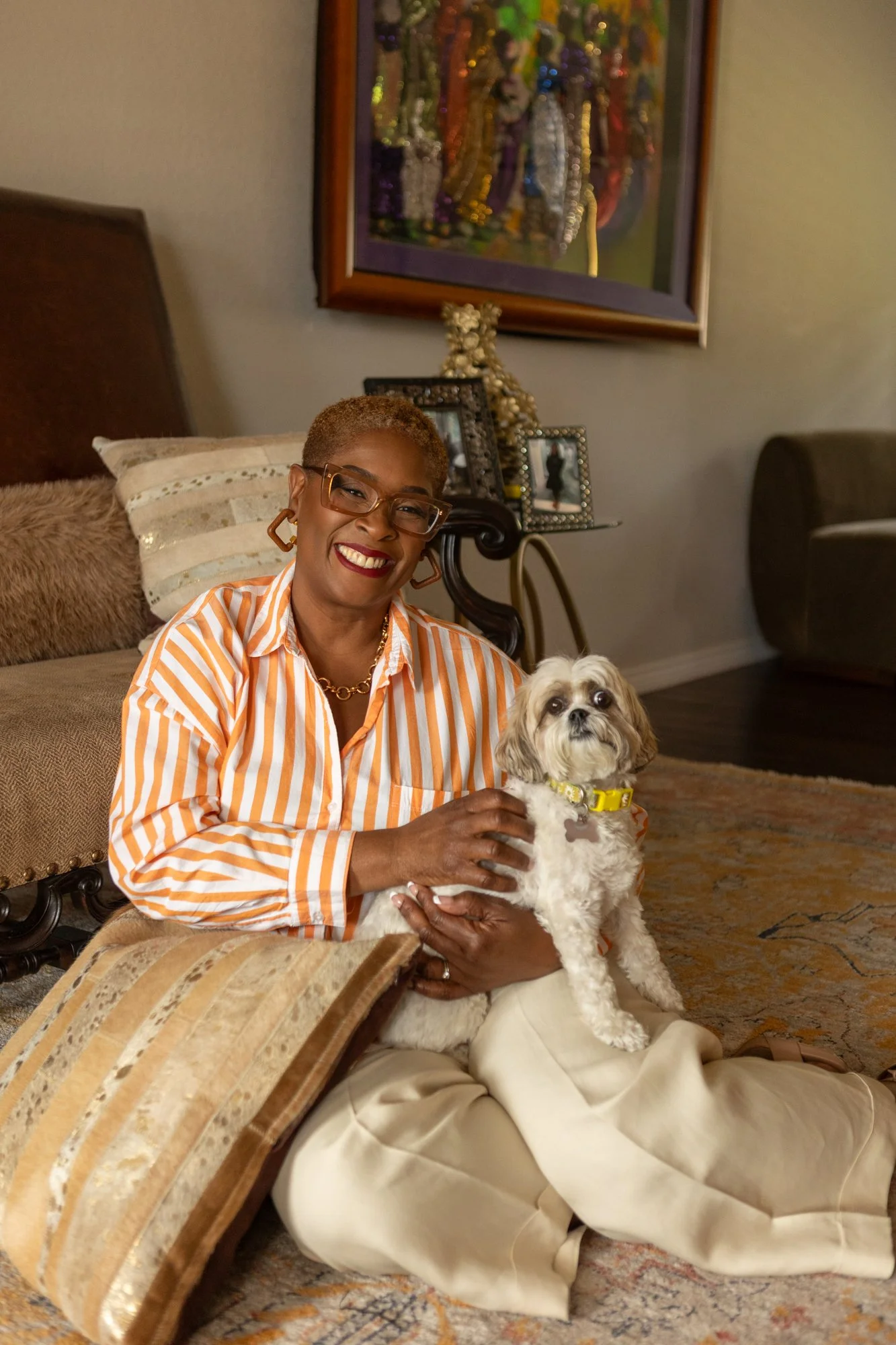 A woman with short, light brown hair and glasses smiling while sitting on the floor in a living room, holding a small Shih Tzu dog with a yellow collar and a bone-shaped tag. She is wearing a striped orange and white shirt.
