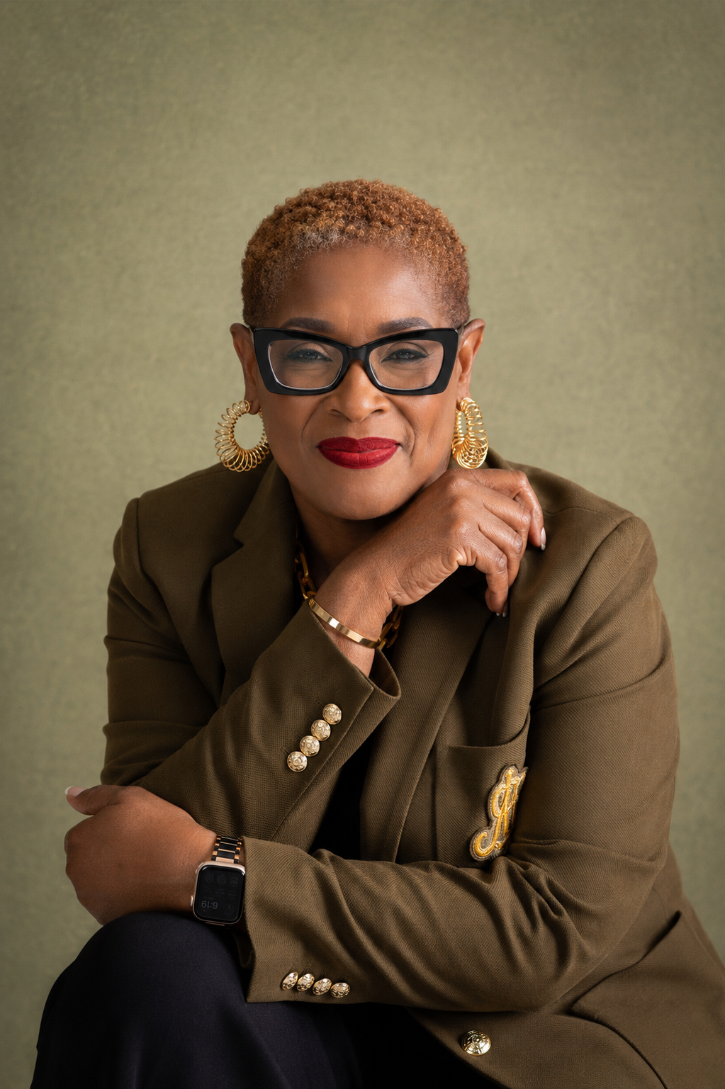 Portrait of a confident woman with short curly hair, wearing black glasses, gold earrings, red lipstick, a gold bracelet, and a brown blazer with gold buttons and embroidery, sitting against a green background.