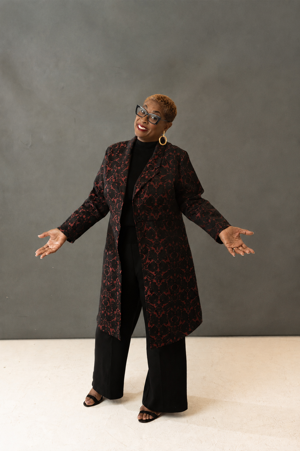 A woman with short, styled hair, wearing glasses, a black top, and a long, dark patterned coat with red accents, standing against a grey background with her arms open and smiling.