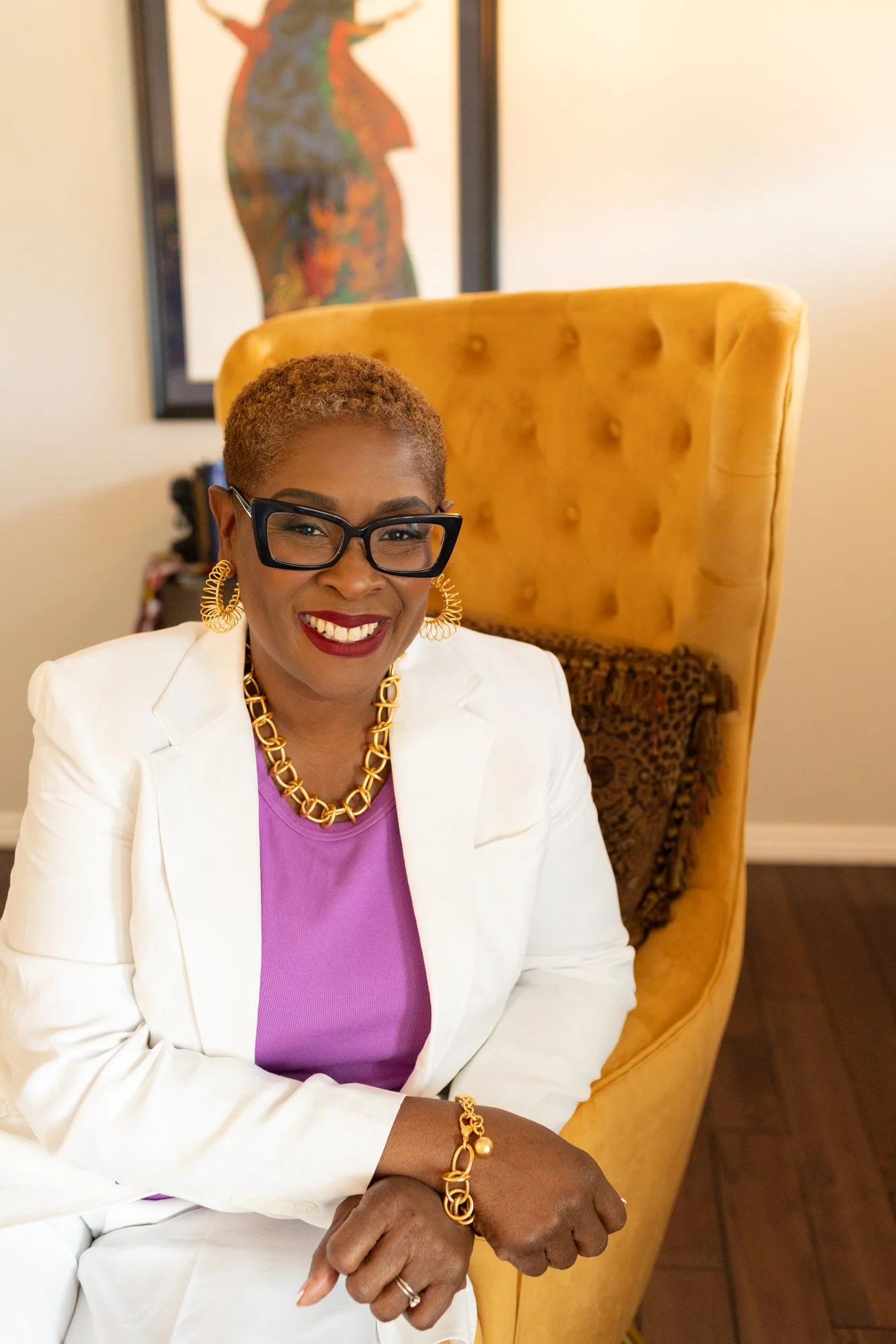 Smiling woman with short curly hair, glasses, red lipstick, wearing gold jewelry, a white blazer, and a purple top, sitting in front of a yellow high-backed chair with a leopard print pillow, with a colorful abstract painting hanging on the wall in the background.