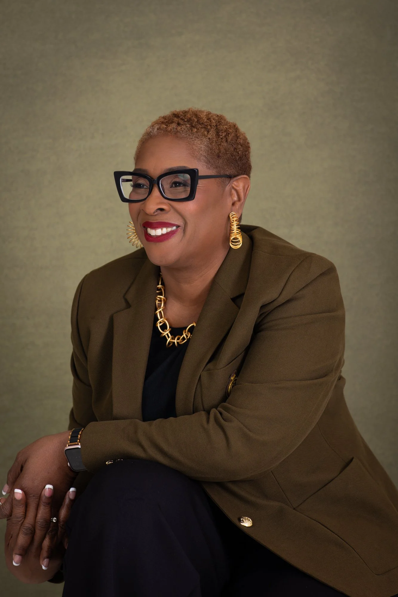 Medium shot of an African American woman with short curly reddish hair, wearing large black glasses, gold earrings, a gold necklace, a watch, and a brown blazer over a black top, sitting with her hands clasped on her knee, smiling, against a plain greenish background.