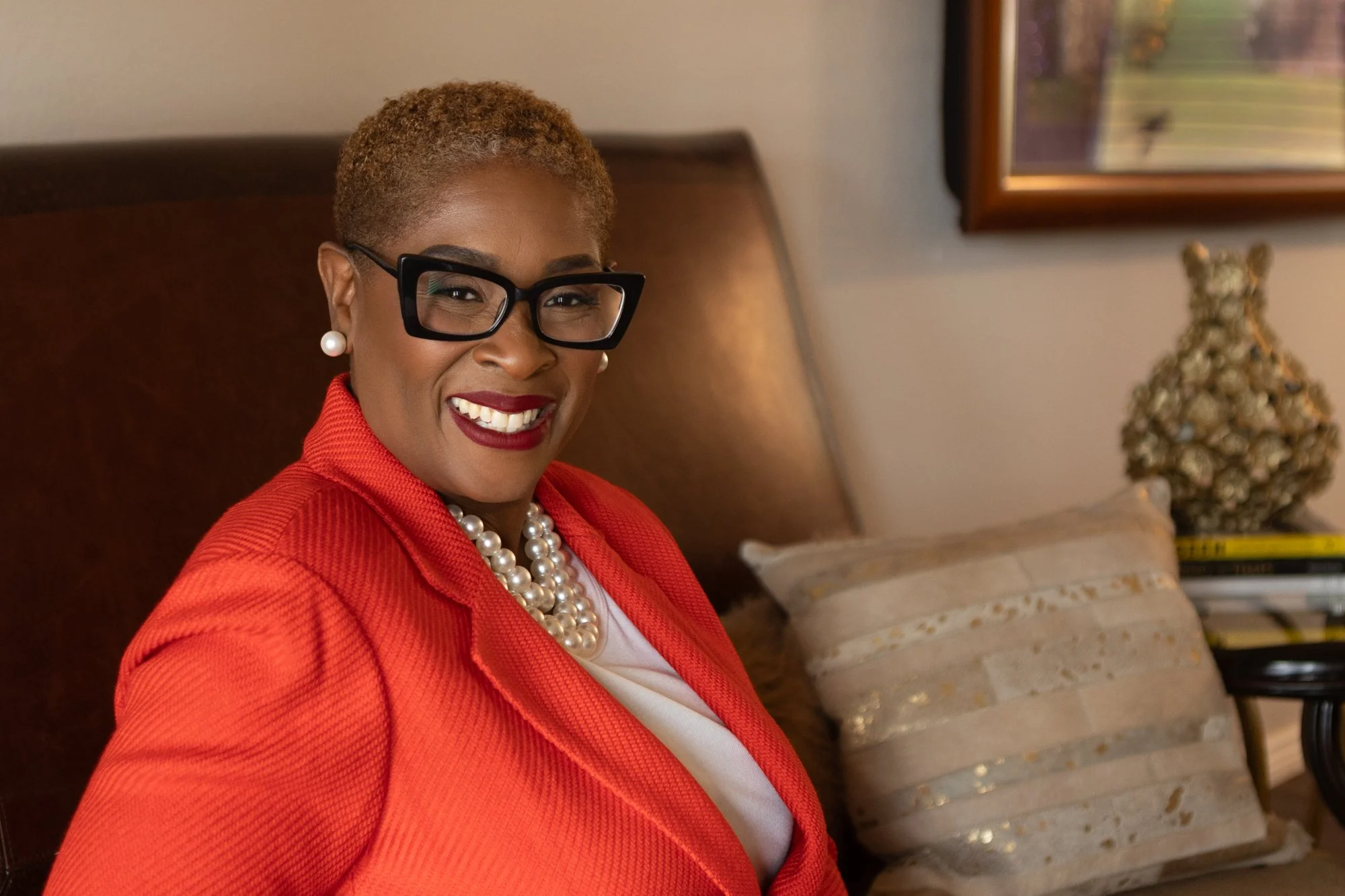 A woman with short, curly, blonde hair and black glasses smiling while sitting on a beige/straw-colored couch in a living room. She is wearing a red blazer, a white top, pearl earrings, and a pearl necklace. Behind her, there is a framed picture on the wall, a ceramic or decorative sculpture on a side table, and some books or magazines stacked.