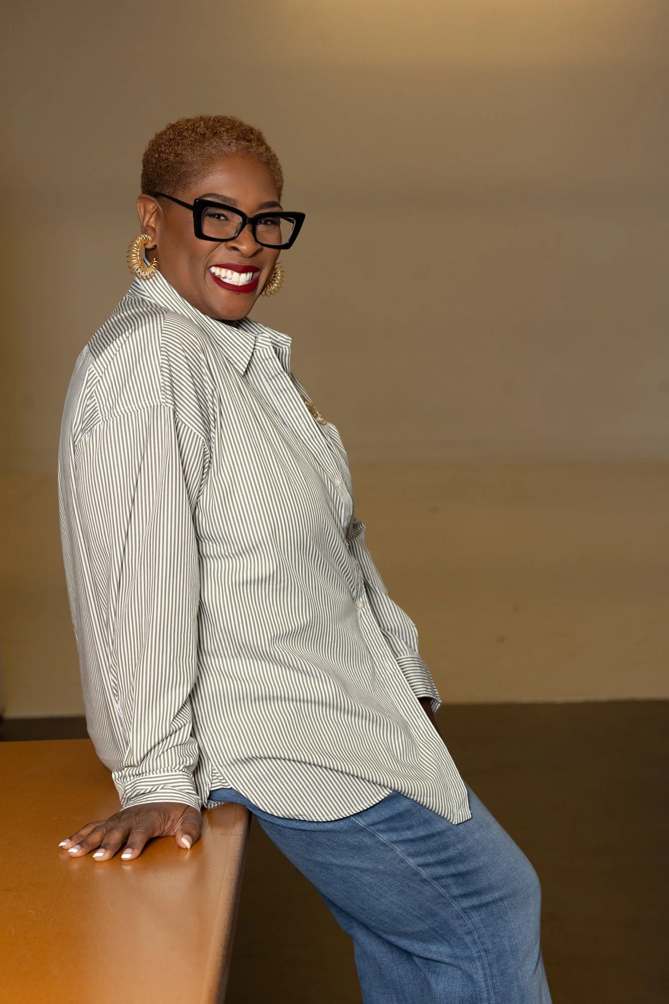 A woman with short, curly hair, wearing glasses, large gold hoop earrings, a striped button-down shirt, and blue jeans, leaning against a wooden surface with a brown wall background, smiling at the camera.