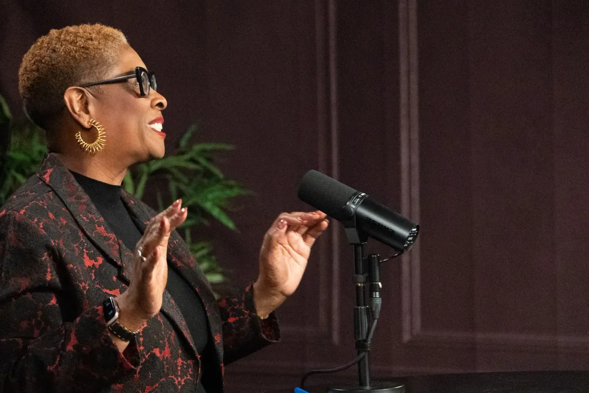 A woman with short curly hair, wearing sunglasses, hoop earrings, a black top, and a floral blazer, speaking into a microphone in a dimly lit room with dark walls and a green plant in the background.