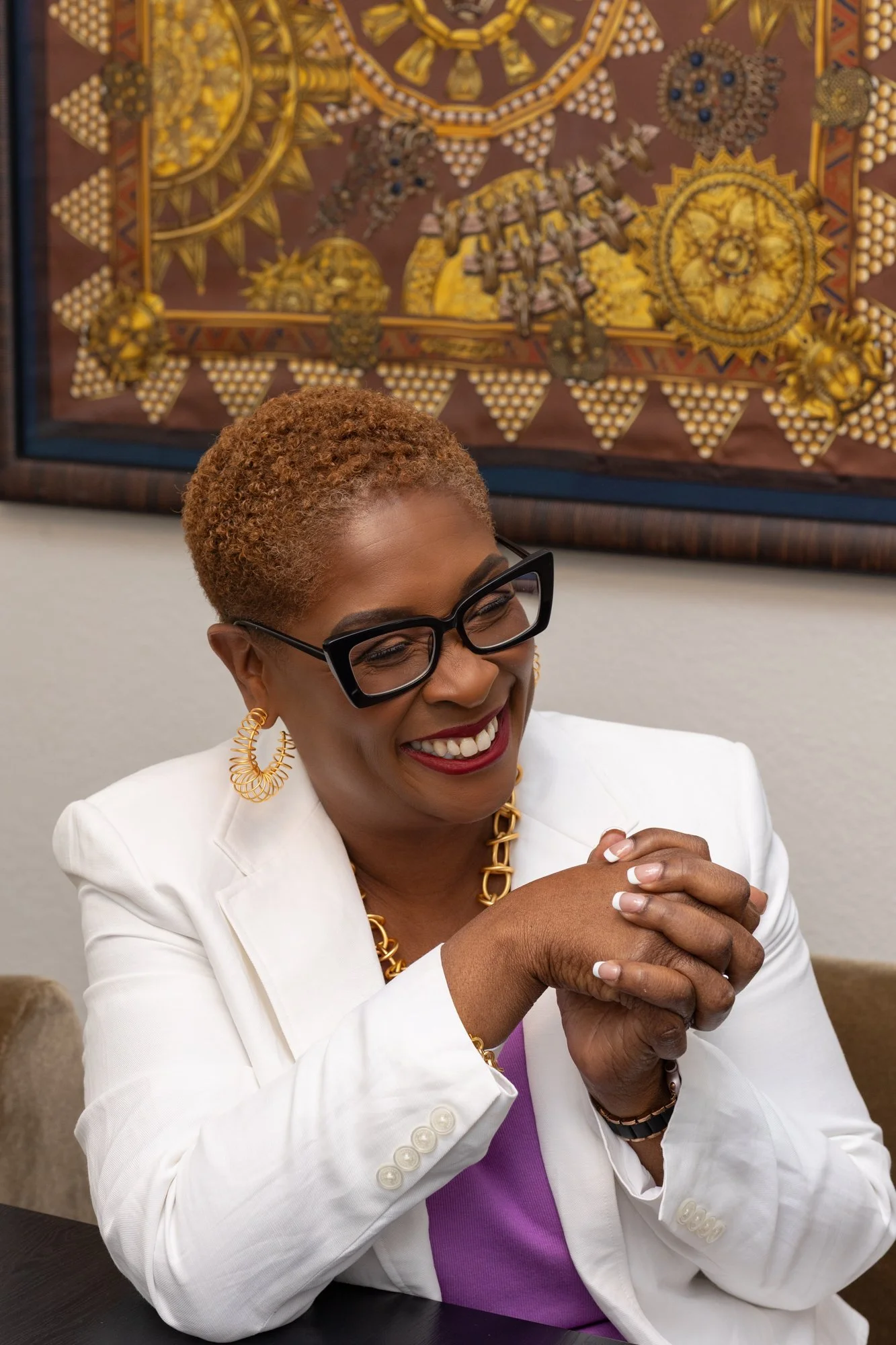 A smiling woman with short curly reddish hair, wearing glasses, gold jewelry, a white blazer, and a purple top, sitting at a table with her hands clasped. Behind her is a colorful decorative artwork on the wall.