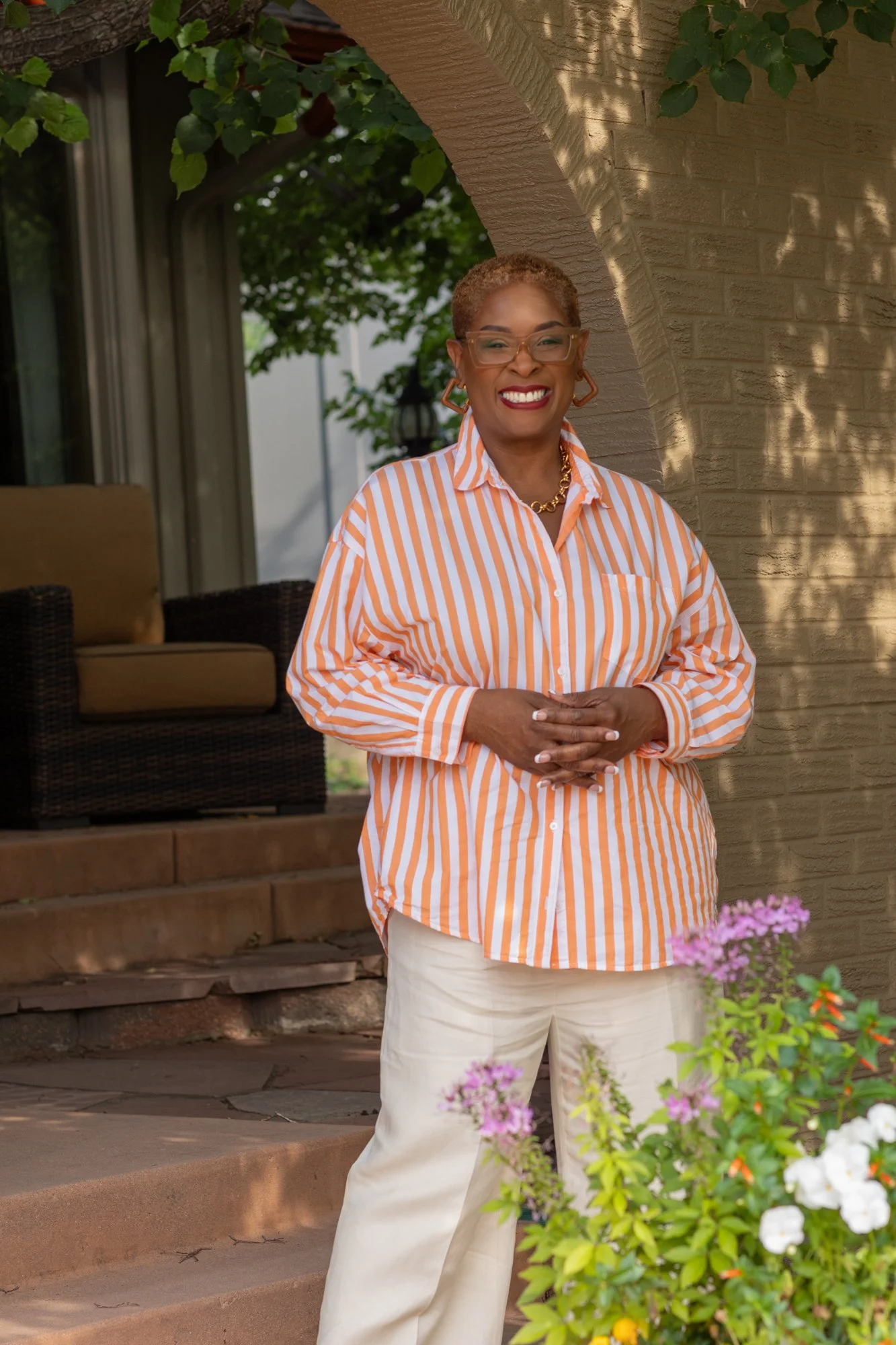 A woman with short curly hair, wearing glasses, a striped orange and white button-up shirt, gold earrings, and a gold necklace, standing outside near a brick archway and flowering plants, smiling with her hands in front of her.