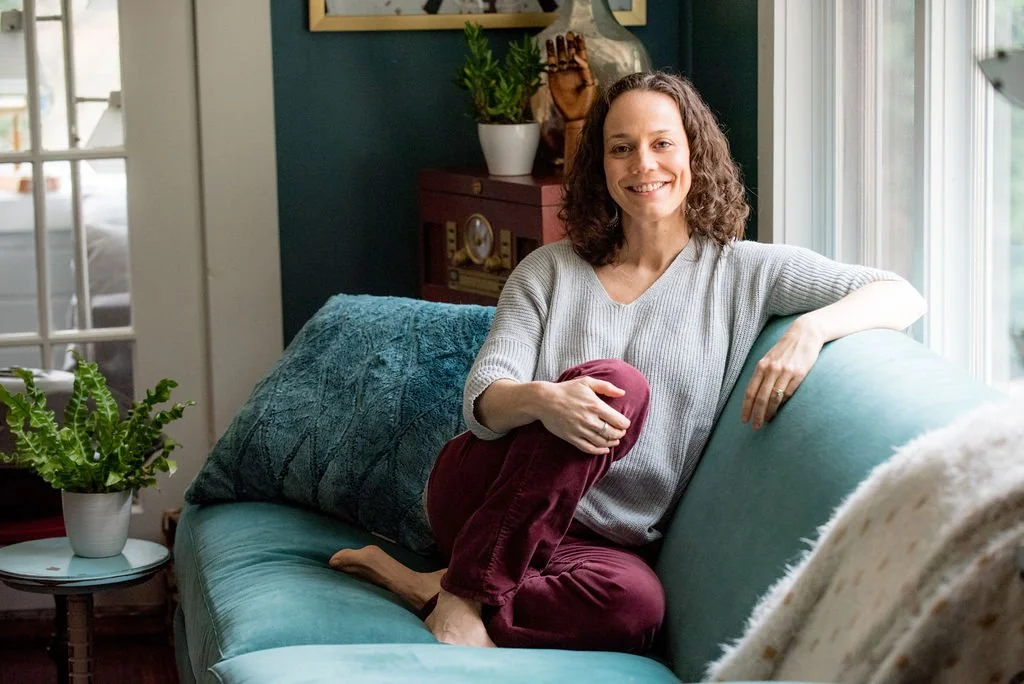 A woman with curly hair smiling and sitting on a teal sofa in a well-lit living room, holding her knee.