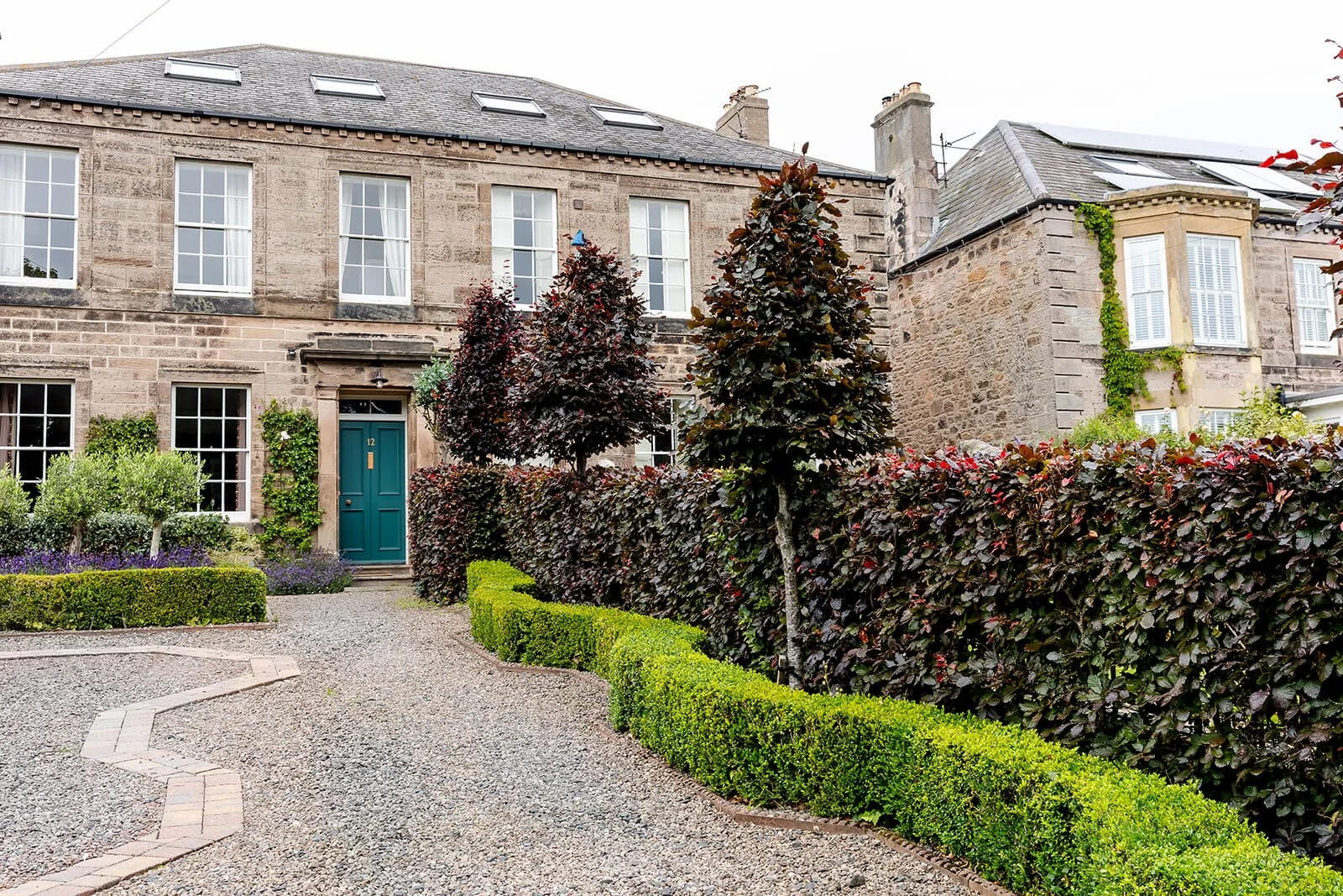 A traditional stone townhouse with a teal front door, surrounded by a manicured garden with shrubs, trees, and a gravel pathway.